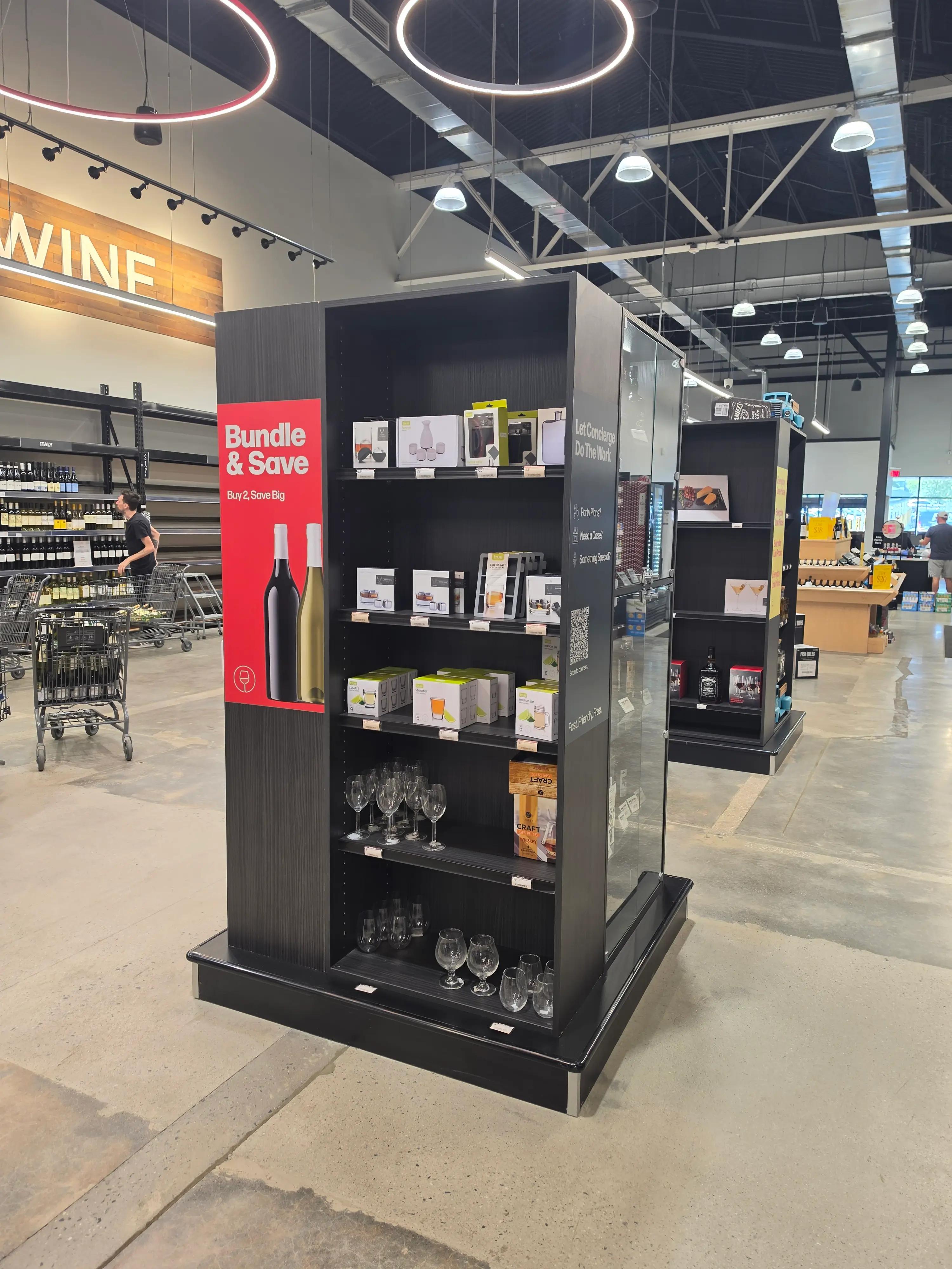 Store interior with a display shelf of wine glasses and bar accessories. A red sign reads "Bundle & Save, Buy 2, Save Big." The background shows a wine section and shopping carts, with modern lighting and high ceilings.