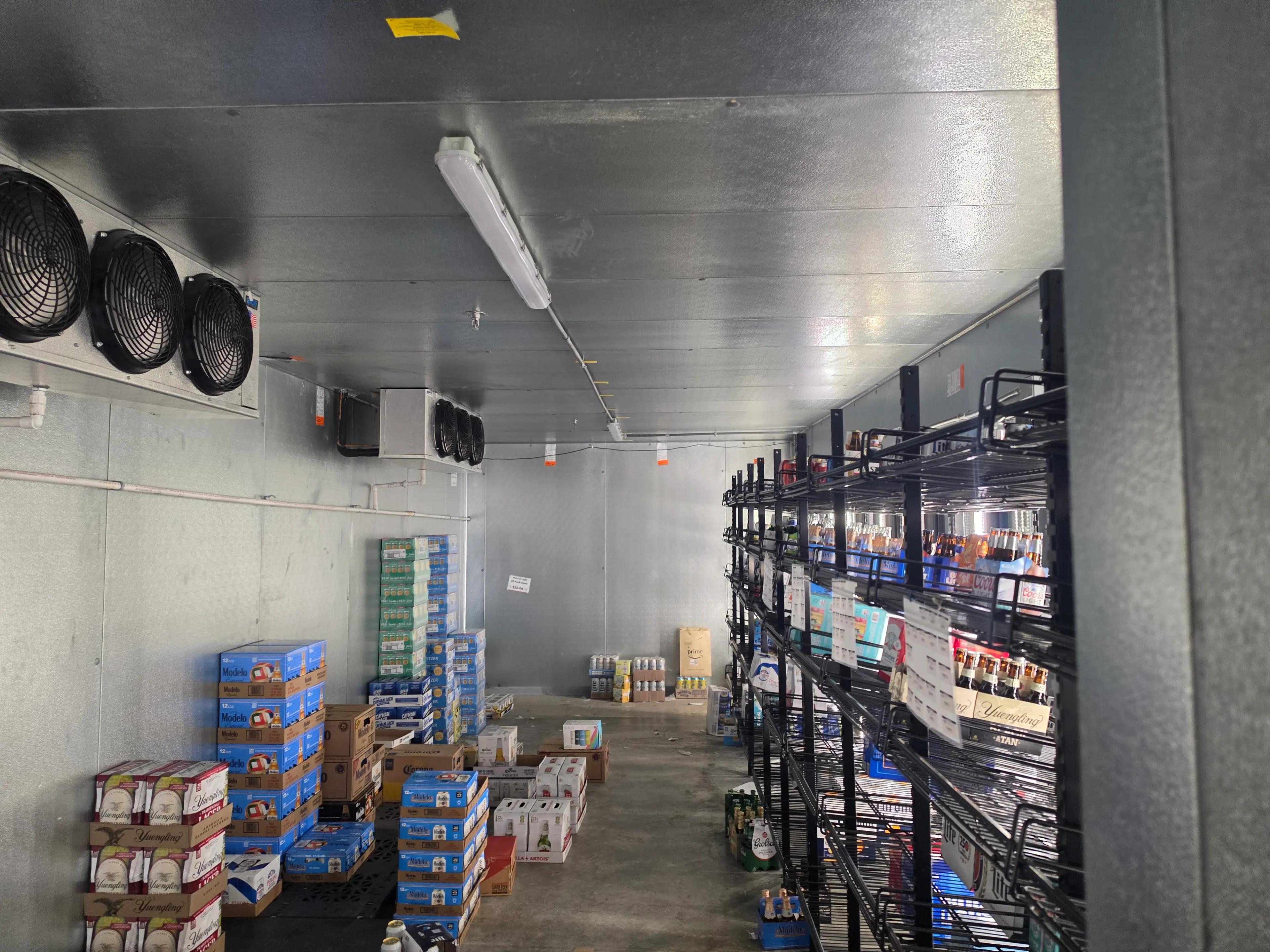 Industrial storage room with metal shelves stocked with various beer and beverage brands, including Coors Light and Modelo. The room features a metallic ceiling with ventilation fans and fluorescent lighting.