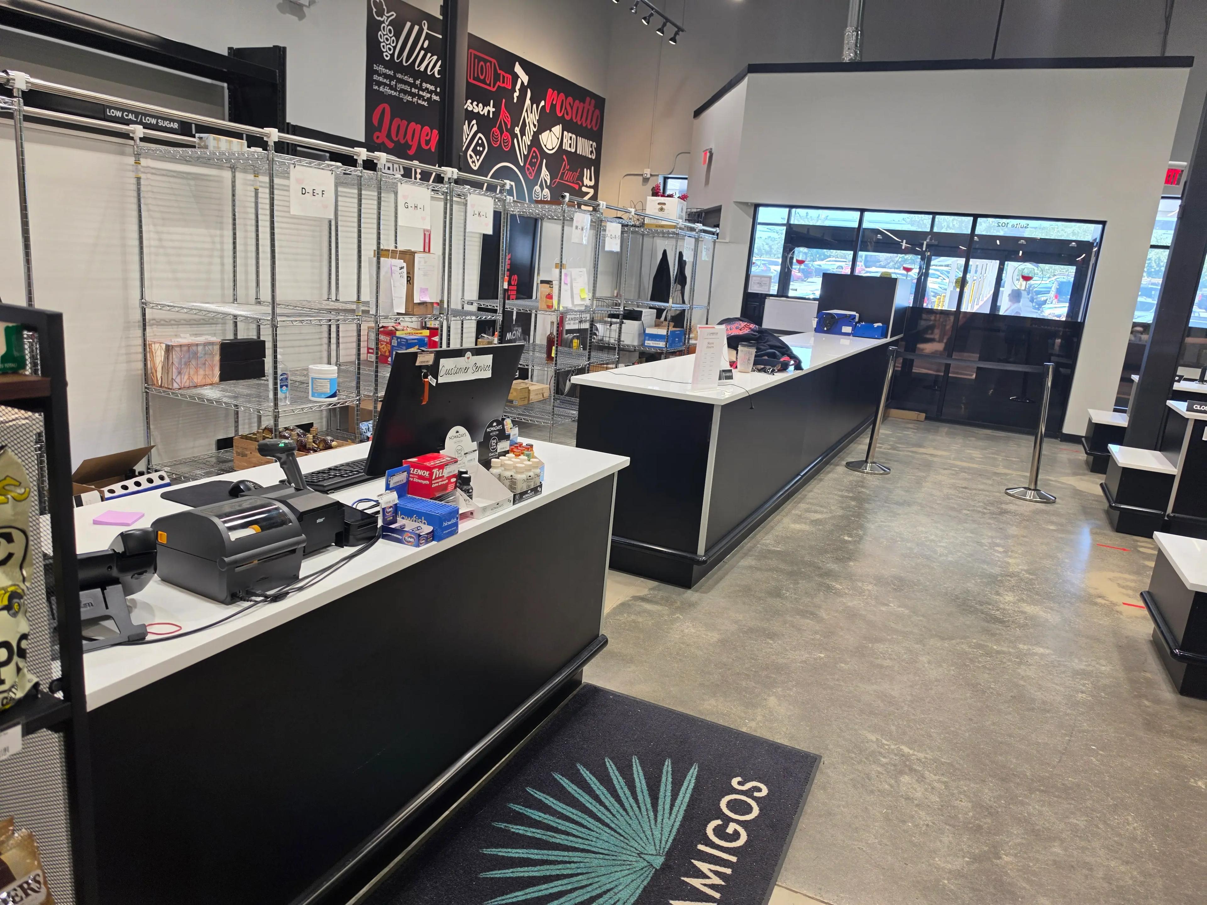 Modern retail store interior with a service counter, shelves labeled for wine and beer, and products like Tums and Tylenol. The floor is polished concrete, and a mat with "Amigos" is near the entrance.