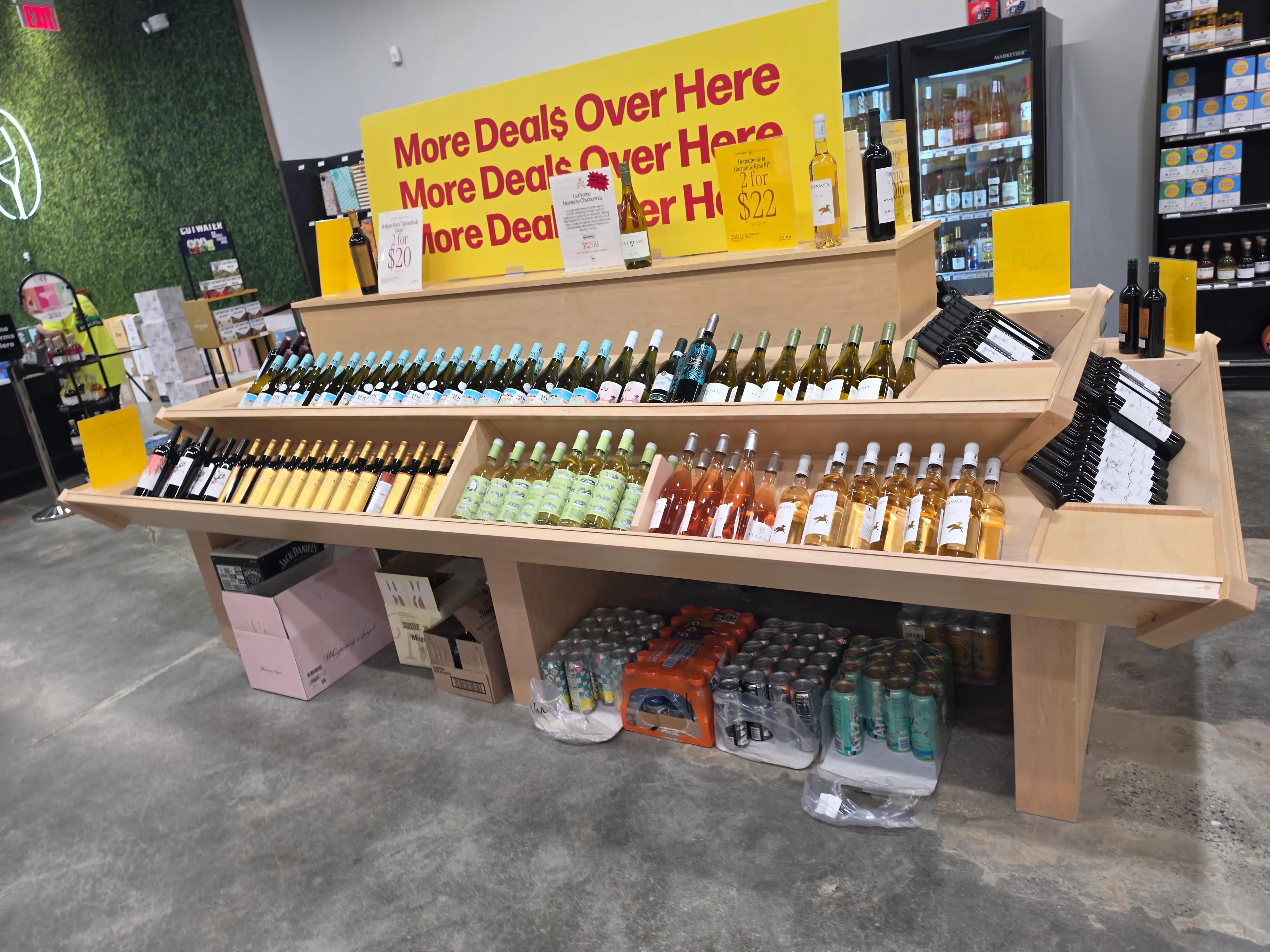 A retail store display featuring various wines and beverages on wooden shelves. A yellow sign reads "More Deals Over Here." Bottles are neatly arranged in rows, with additional drinks stored below.