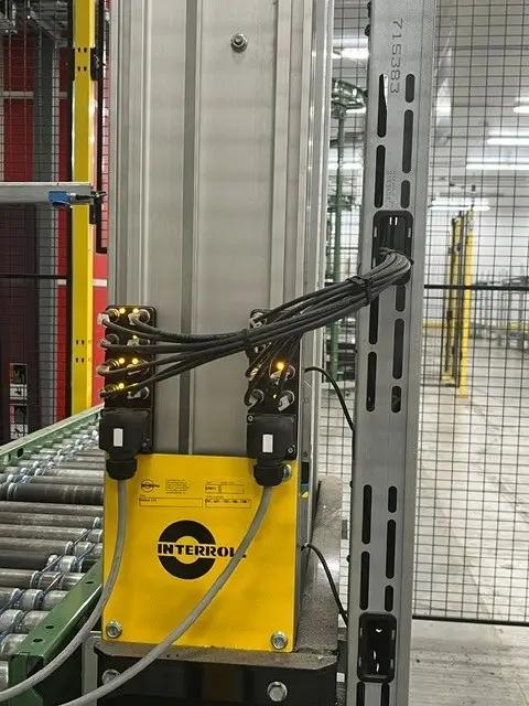 Industrial equipment featuring a yellow Interroll component and wiring, integrated into a conveyor system within a warehouse environment.