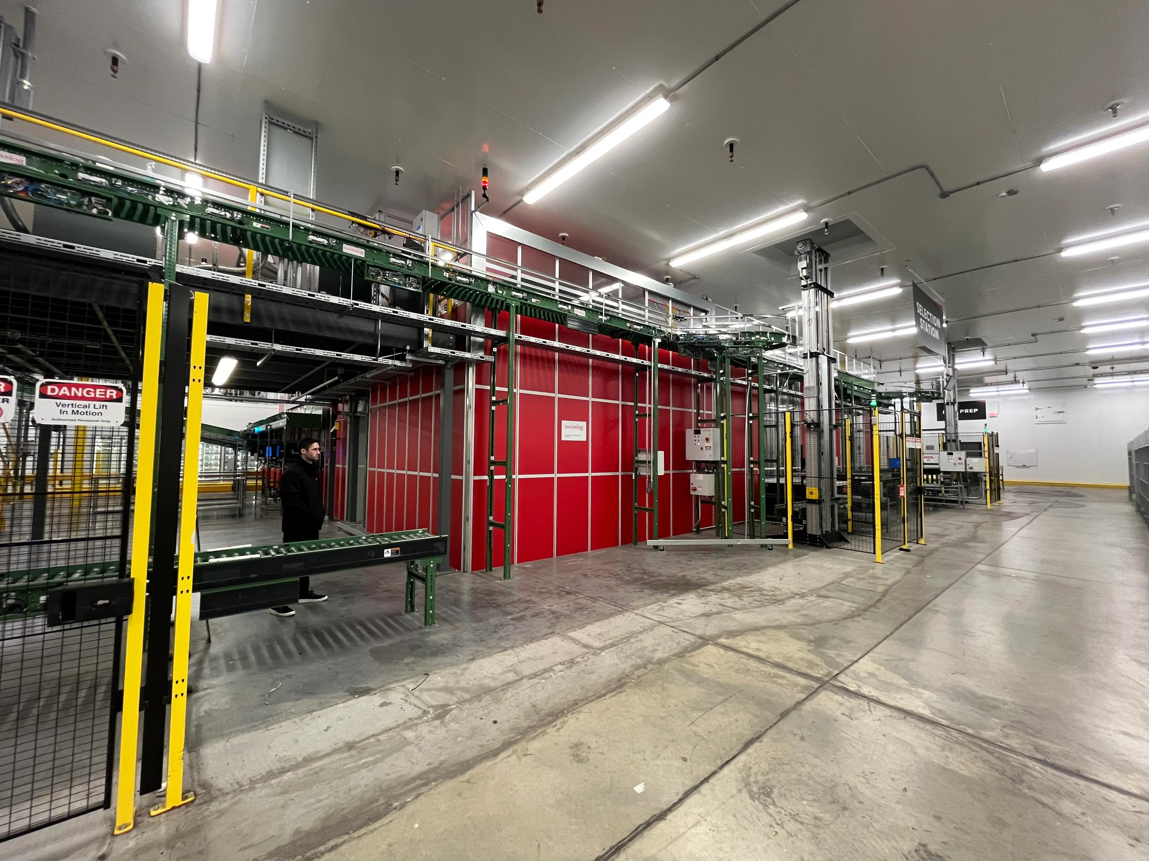 Industrial facility with a red and gray vertical lift system labeled "DANGER Vertical Lift In Motion" and "SELECTION STATION." A person stands nearby in a well-lit area with overhead lights.