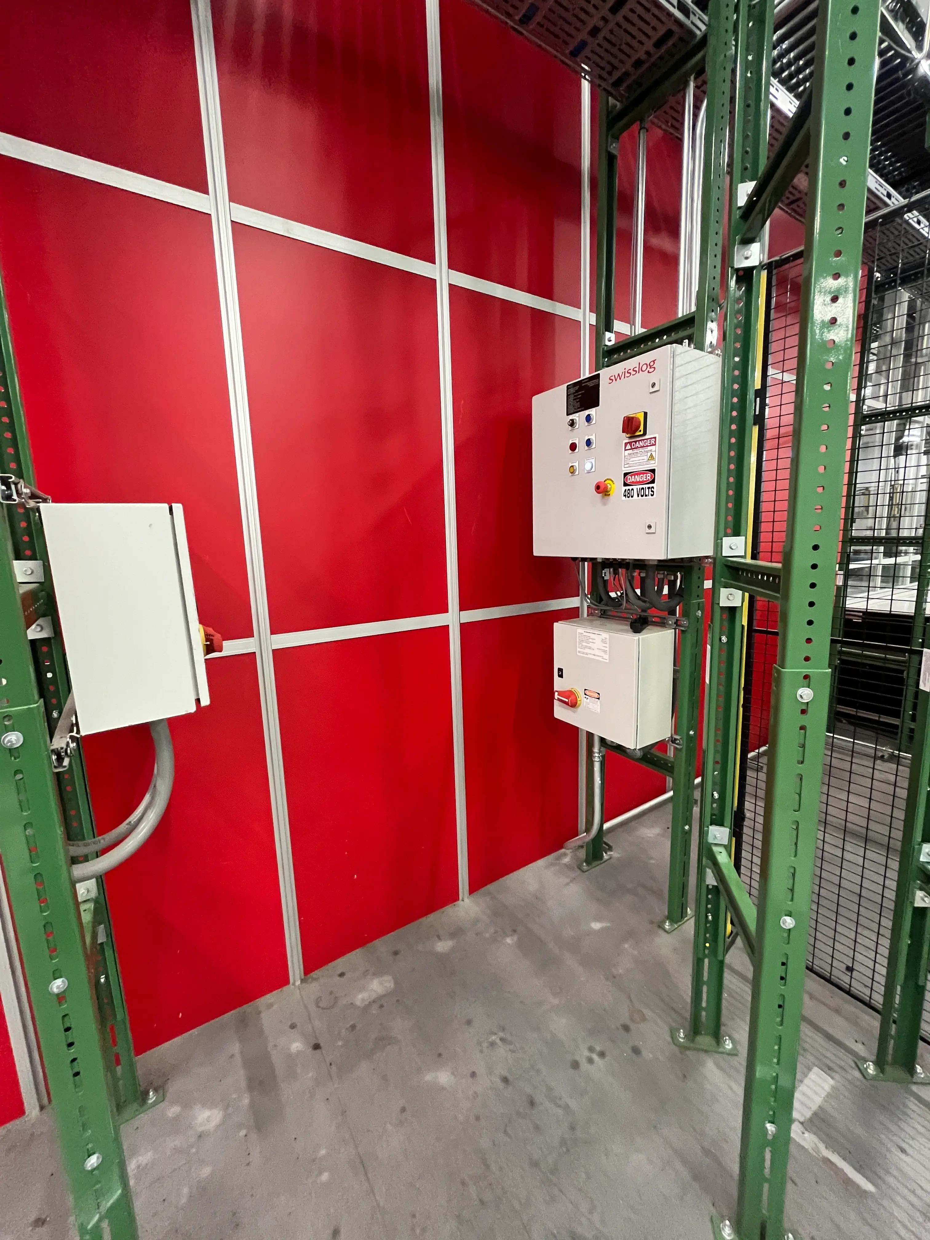 Electrical control panel labeled "swisslog" on a green metal frame against a red wall, with warning labels indicating "DANGER" and "480 VOLTS." Part of a Mid America project.