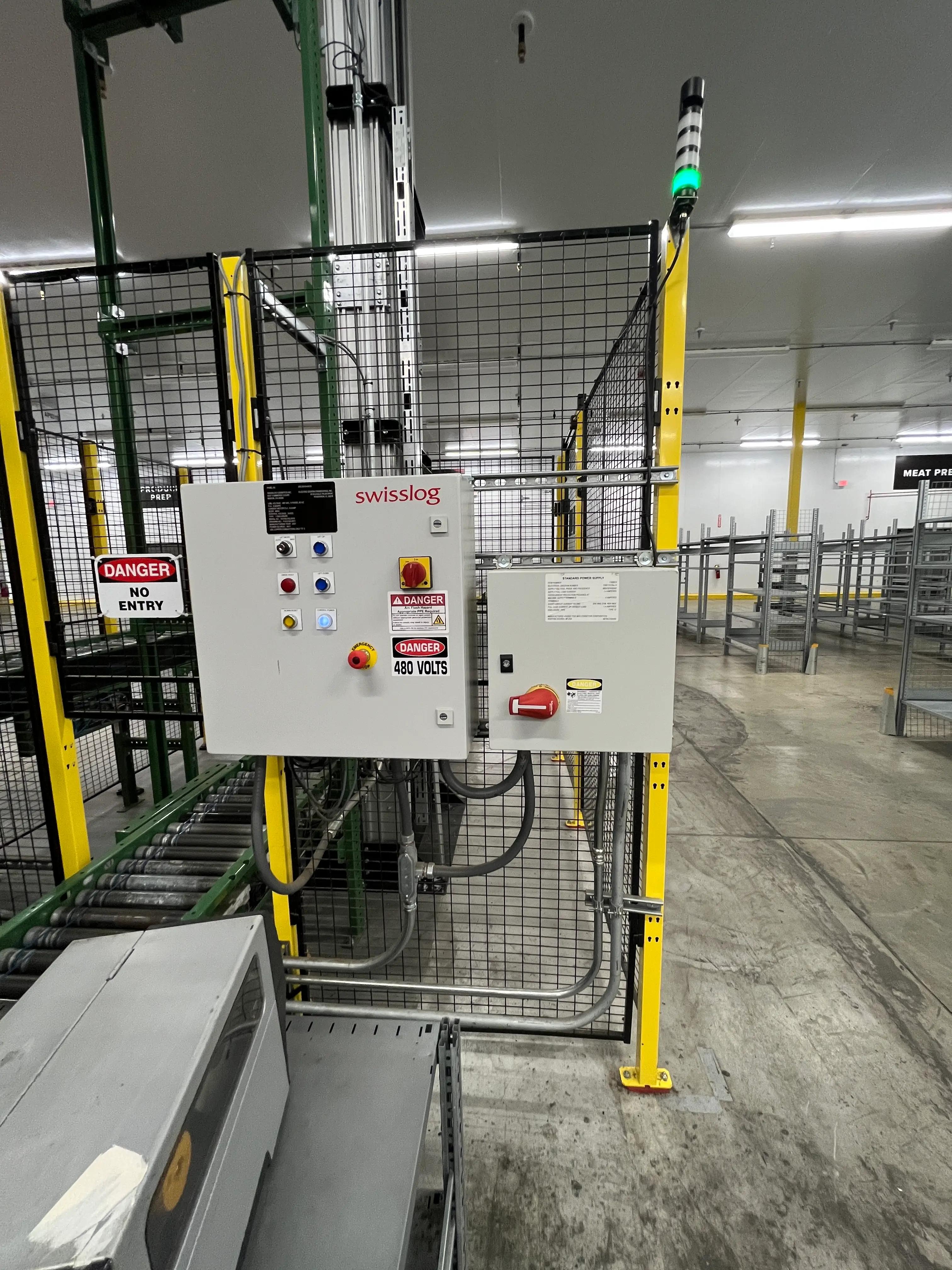 Industrial area featuring a Swisslog control panel with "Danger 480 Volts" and "No Entry" signs, surrounded by metal fencing and conveyor systems, indicating a high-voltage work environment.