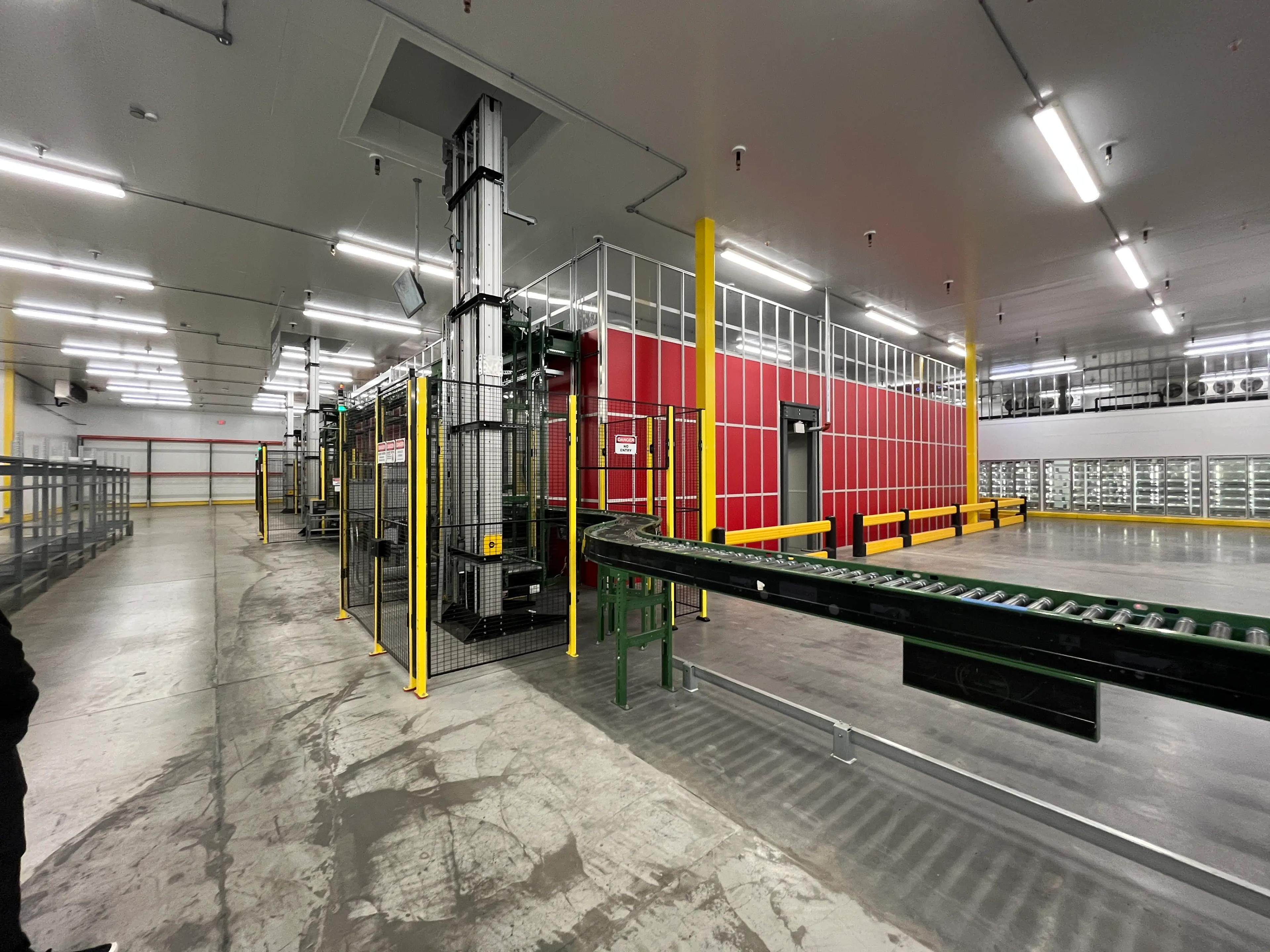 An industrial facility interior featuring conveyor belts, metal structures, and a red and white partitioned area. Overhead lighting brightens the space, with visible safety barriers.