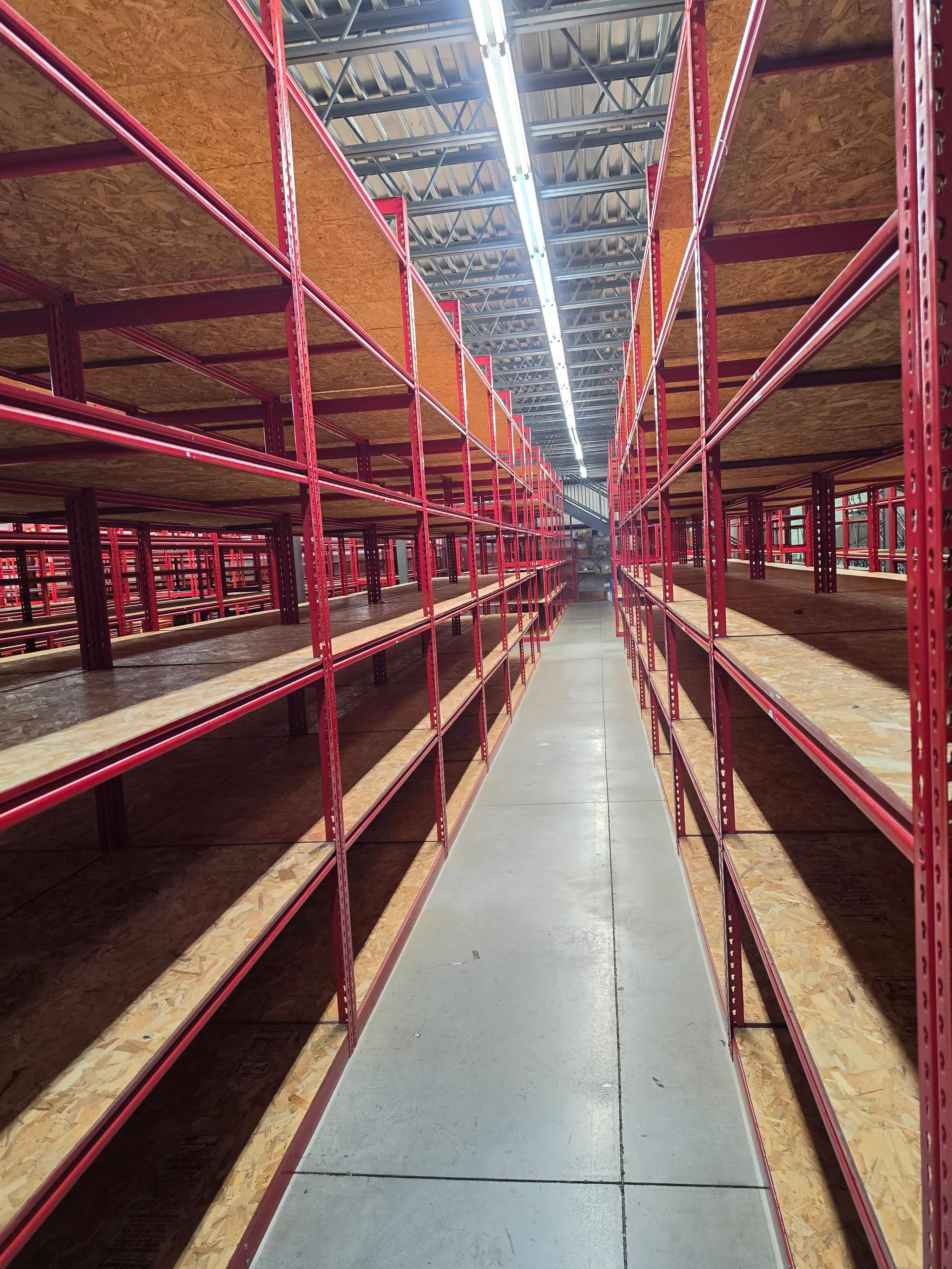 Long warehouse aisle with tall red metal shelving units and wood surfaces under industrial lighting.