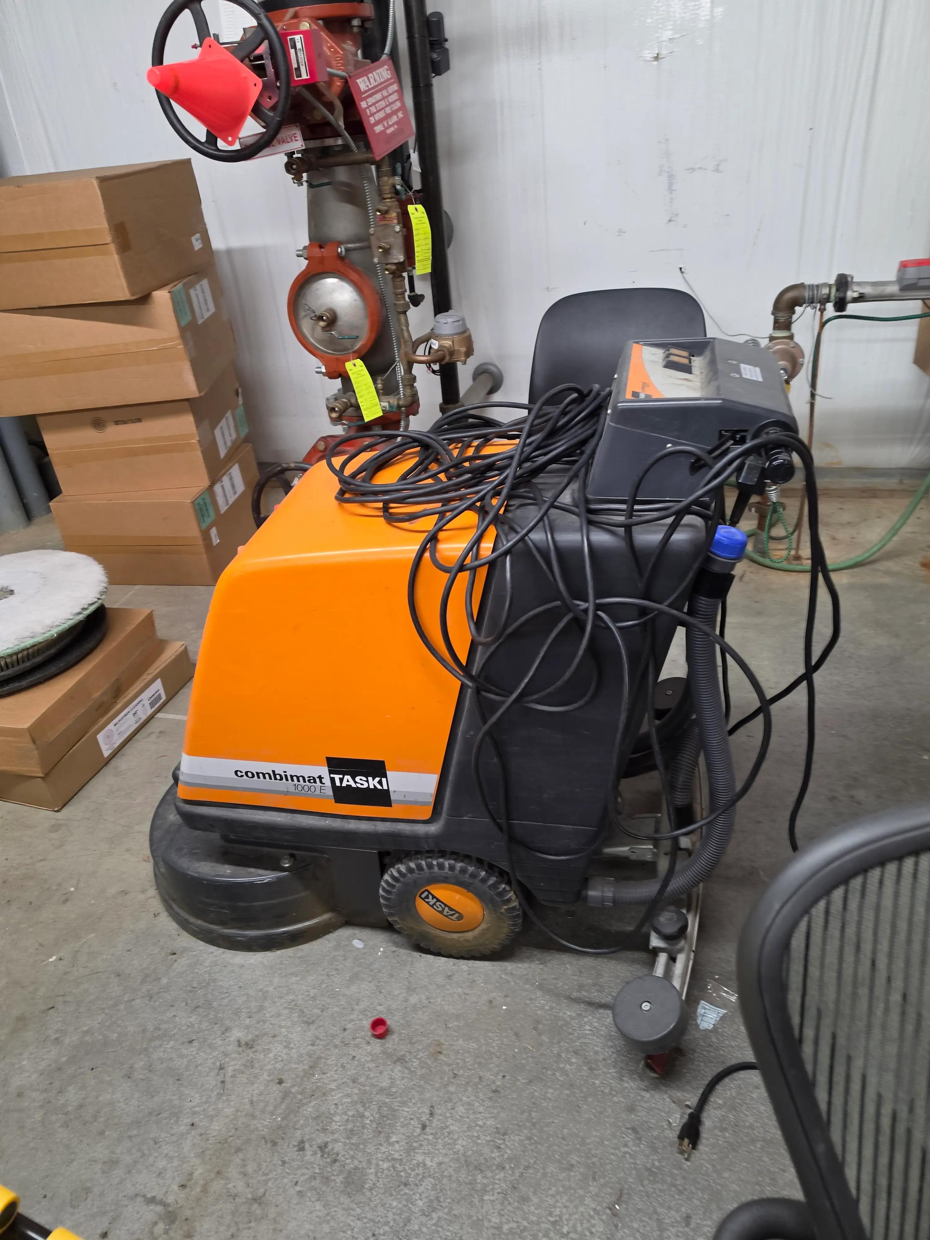 An orange and black floor cleaning machine labeled "combinat TASKI 1000 E" is situated in a storage area, surrounded by boxes and a fire department valve.