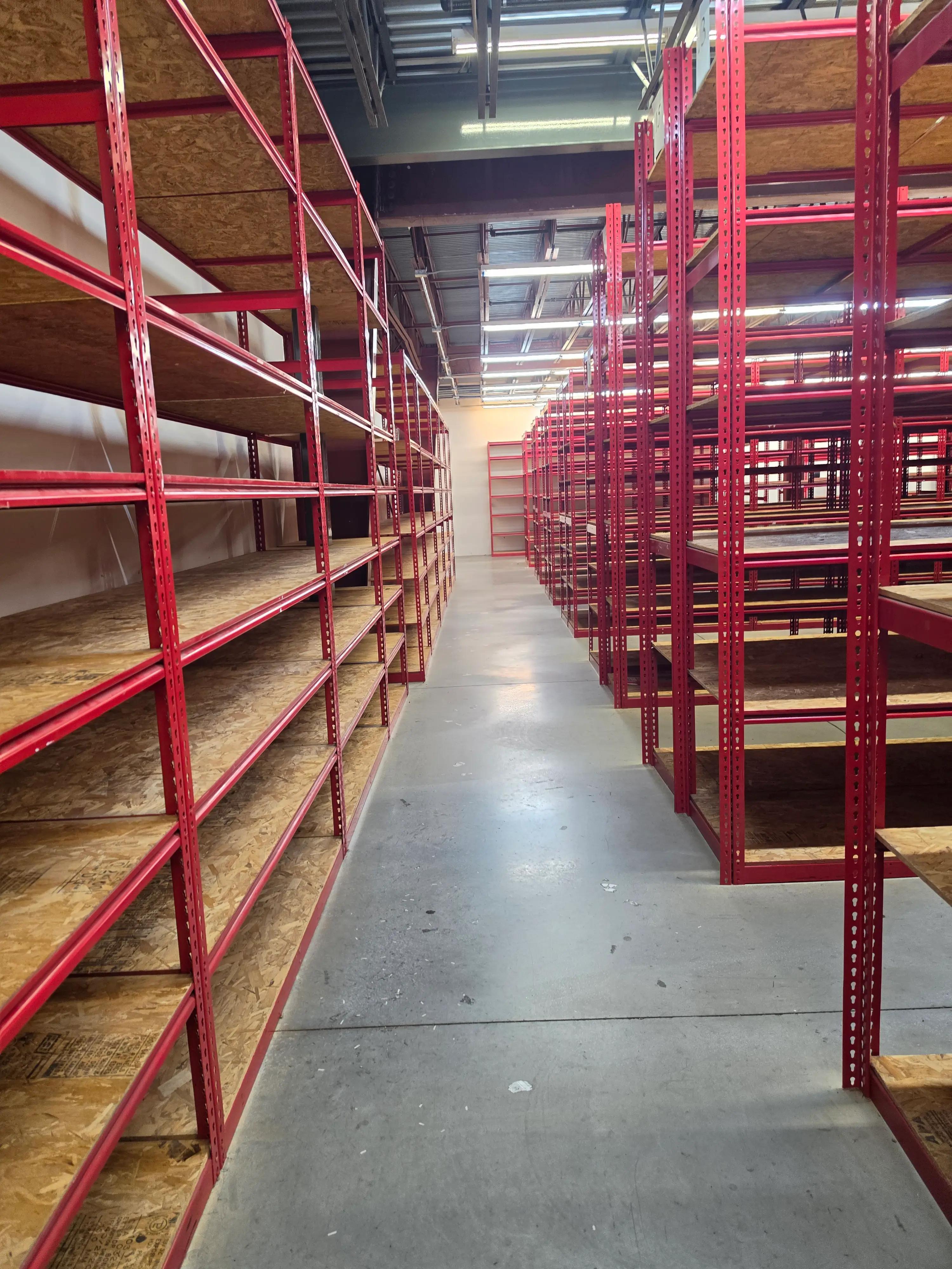 Warehouse aisle with red metal shelving units on both sides, featuring multiple levels of wooden shelves. The floor is concrete, and the ceiling has exposed beams and fluorescent lighting.