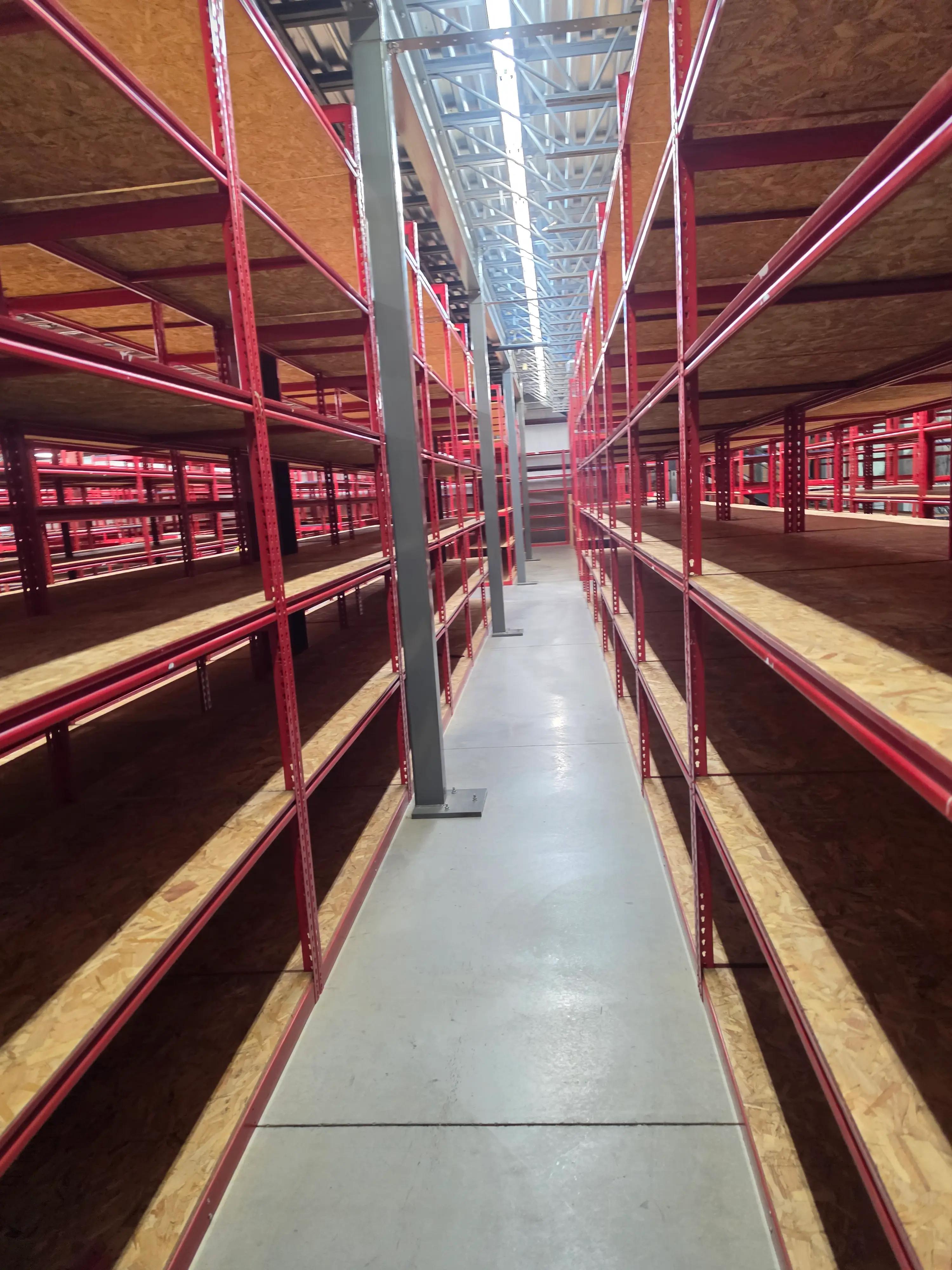 Warehouse aisle with red metal shelving units on both sides, wooden shelves, exposed metal beams on the ceiling, and fluorescent lighting.