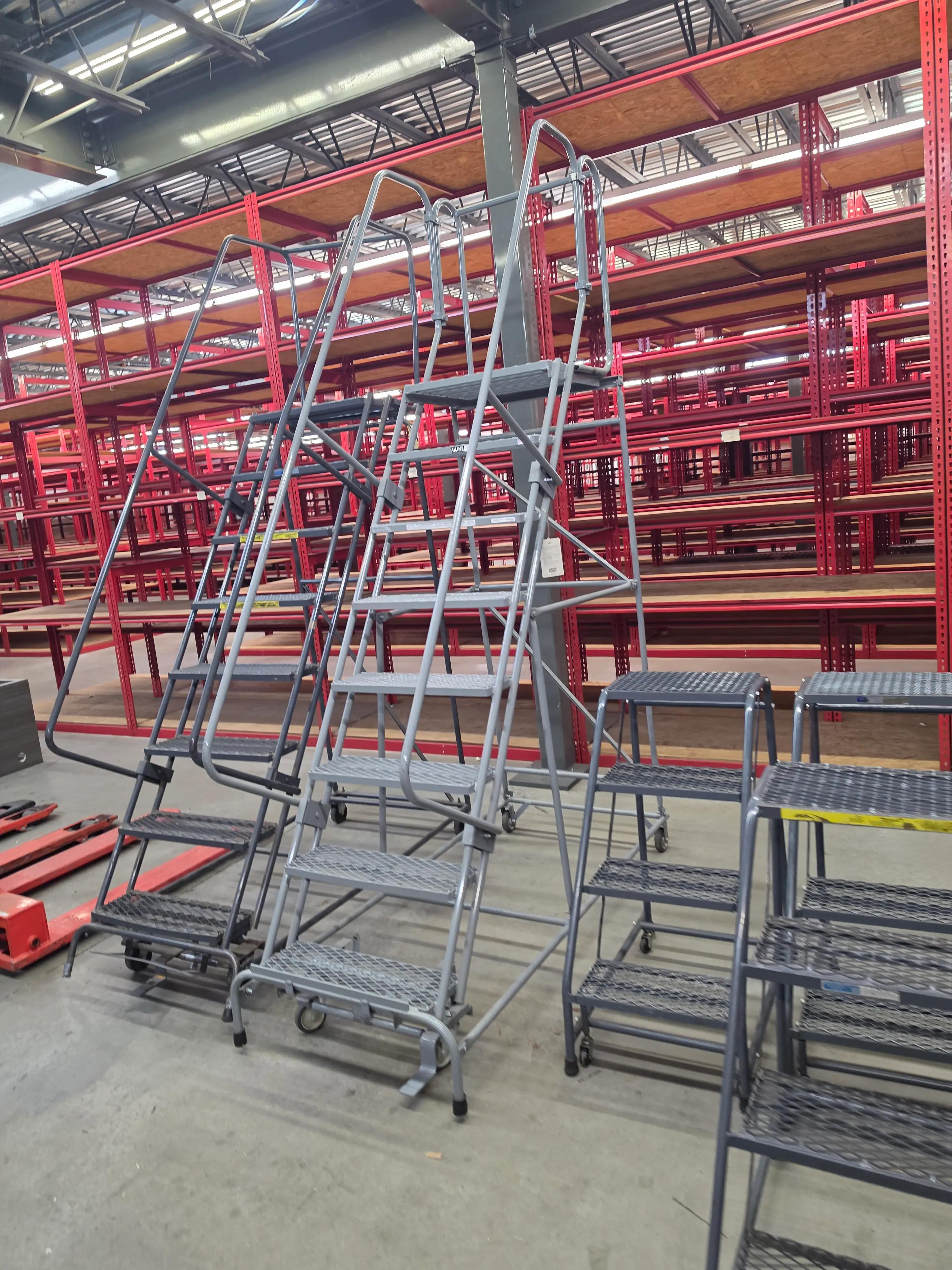 Warehouse interior featuring red metal shelving and several gray rolling ladders on a concrete floor.