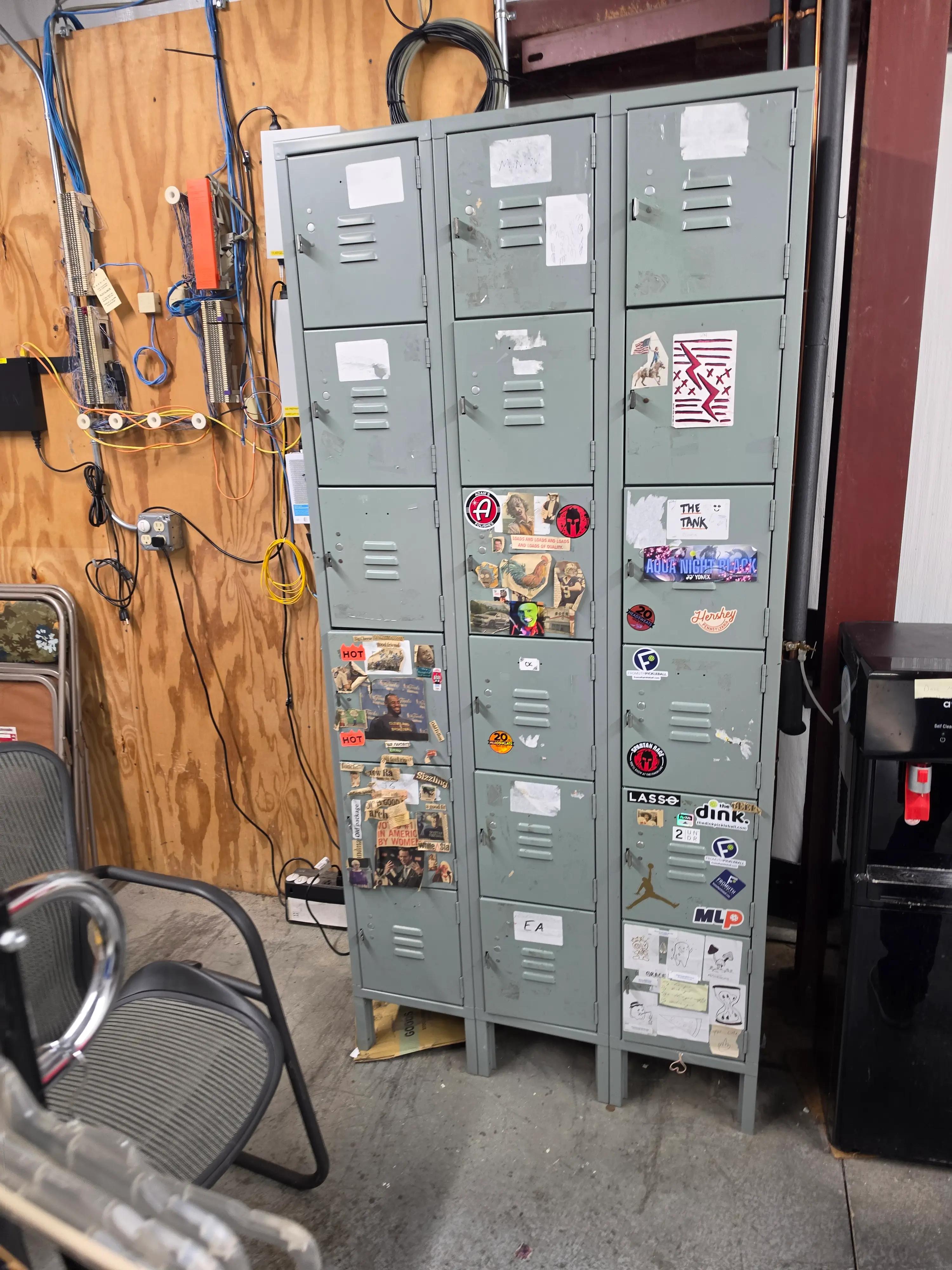 Gray metal lockers adorned with assorted stickers and labels in a workshop setting, featuring visible wiring and a wooden wall. A chair and some equipment are positioned nearby.