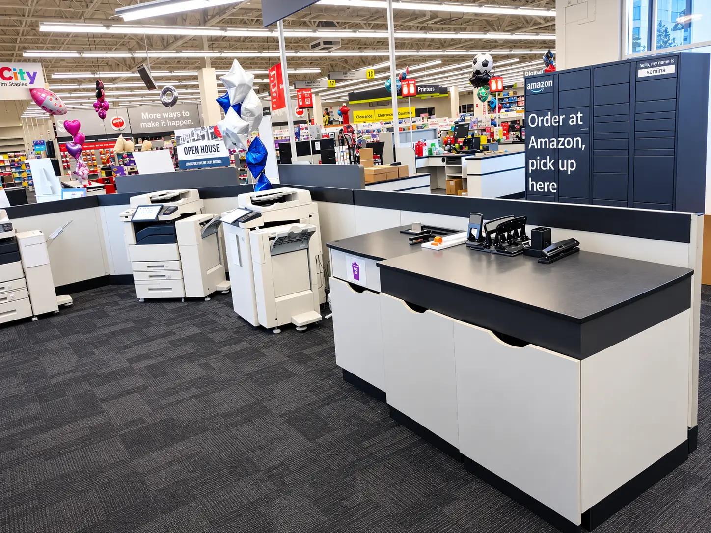 Store interior with a printing station, printers, and office supplies. Signs for Amazon order pickup and clearance sales are visible. Balloons and various store sections are in the background.