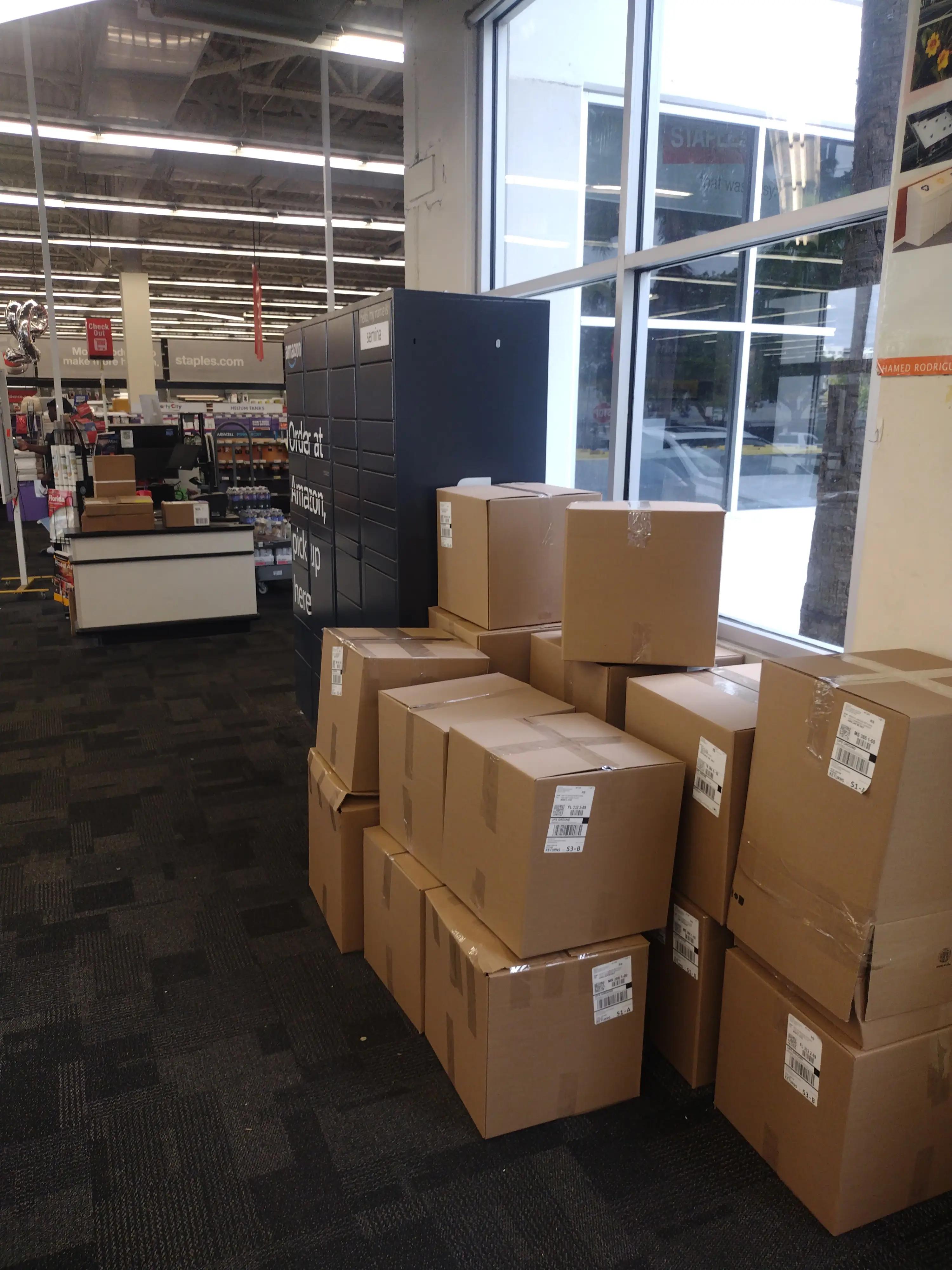 Stacks of cardboard boxes are piled near a window inside a store, with a black Amazon locker nearby. The store features a visible checkout area and various products on display.