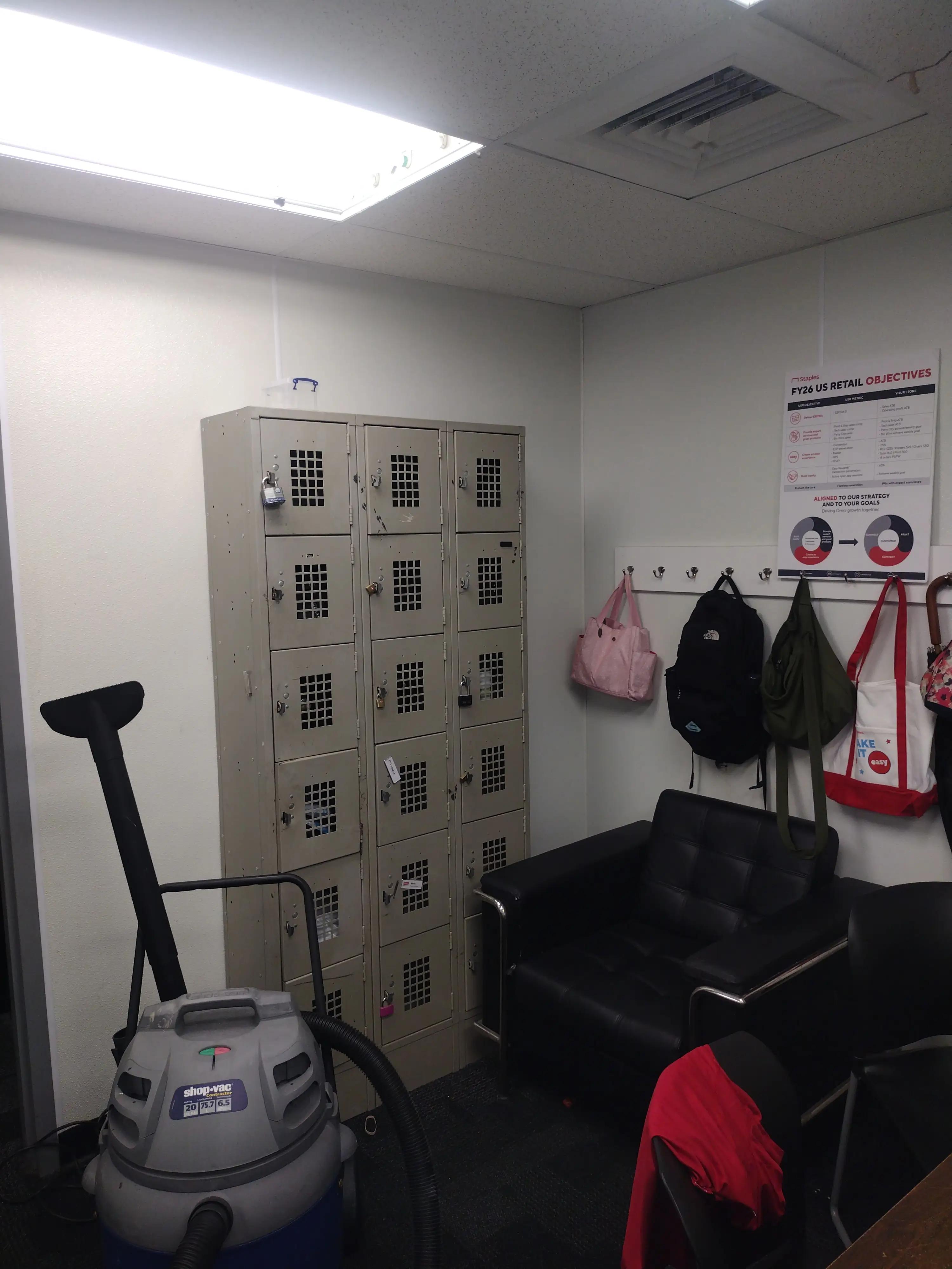 Break room with lockers, a black couch, a shop-vac, and a wall poster detailing FY26 US retail objectives. Backpacks and bags hang on hooks, and a red jacket is draped over a chair.