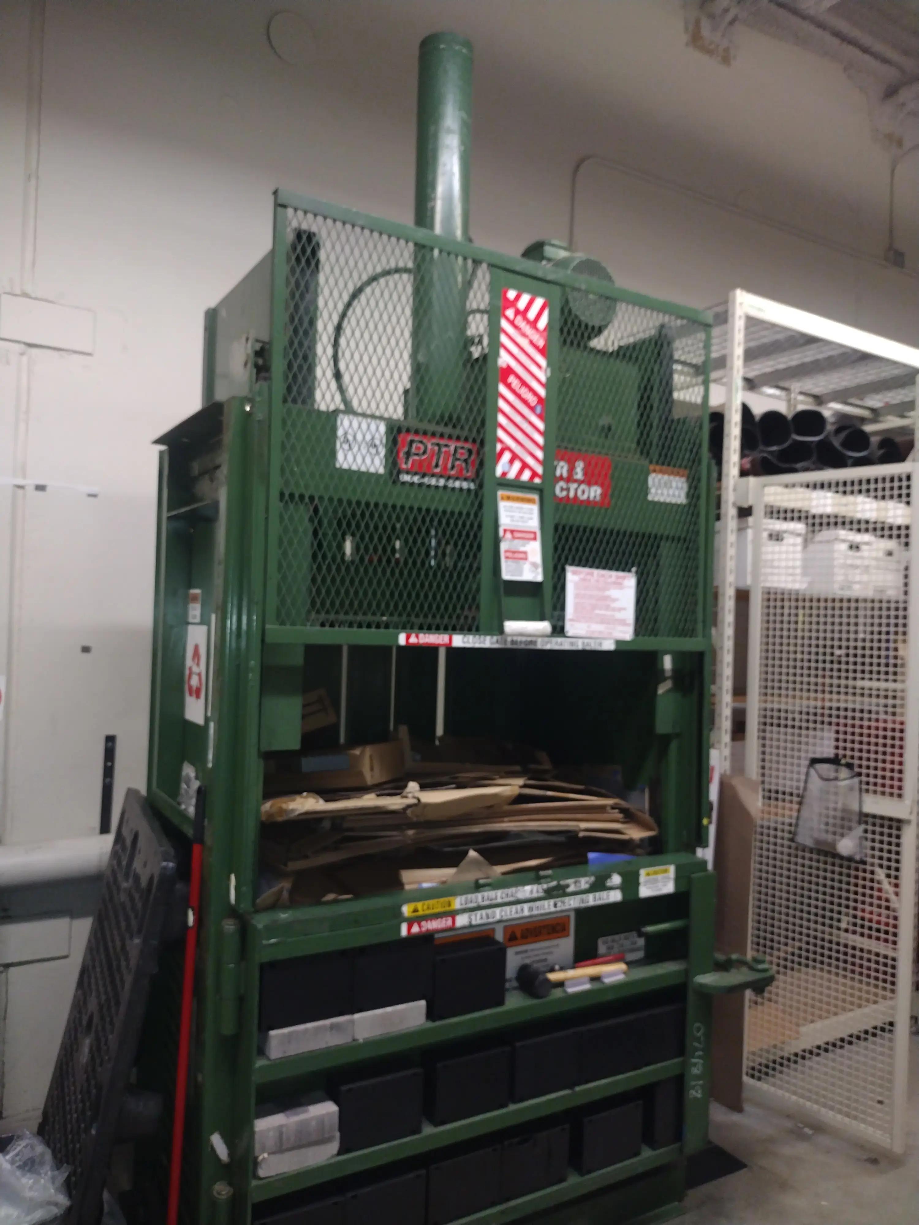 A large green industrial compactor labeled "PTR Baler & Compactor" with cardboard boxes inside, situated in a warehouse with metal shelving and pipes in the background. Safety warnings are visible on the machine.