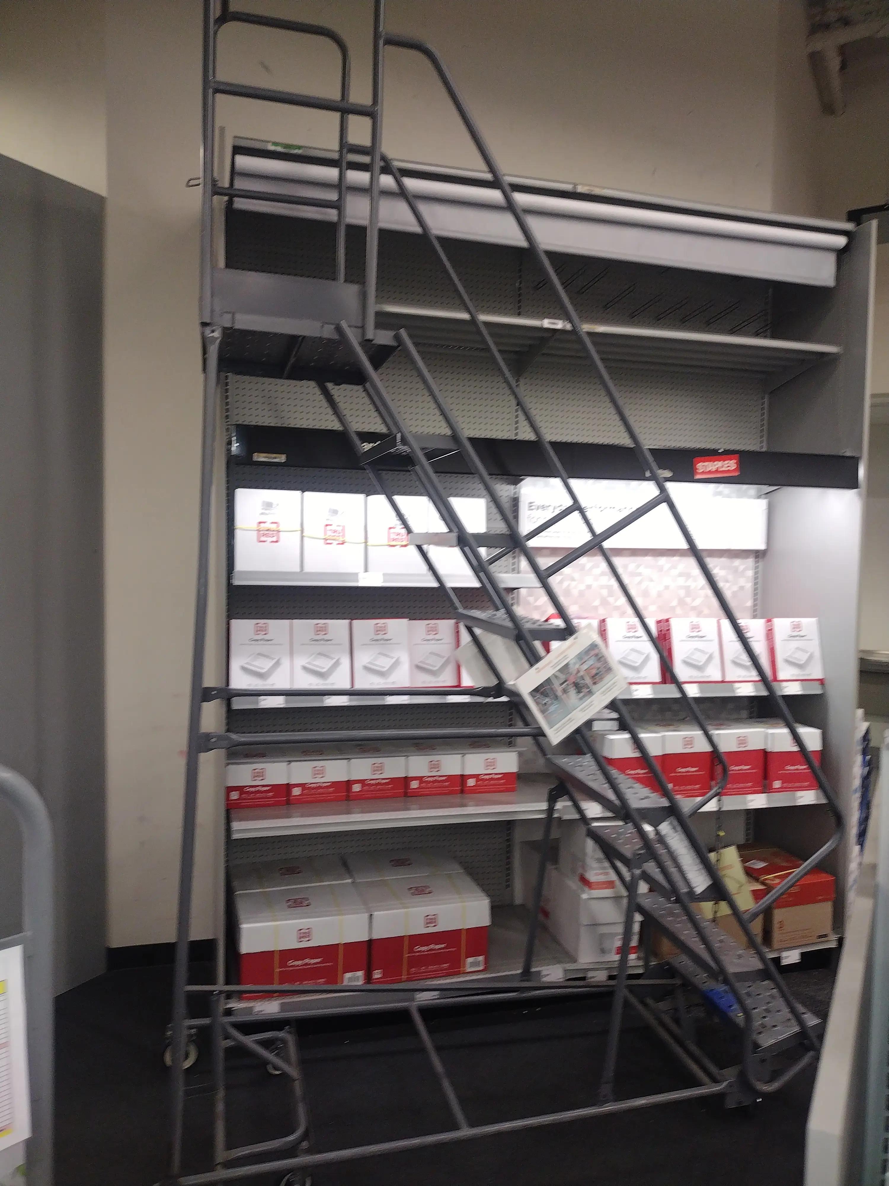 A metal rolling ladder in front of shelves stocked with boxes of paper products at a Staples store, with various office supplies visible and a sign attached to the ladder.