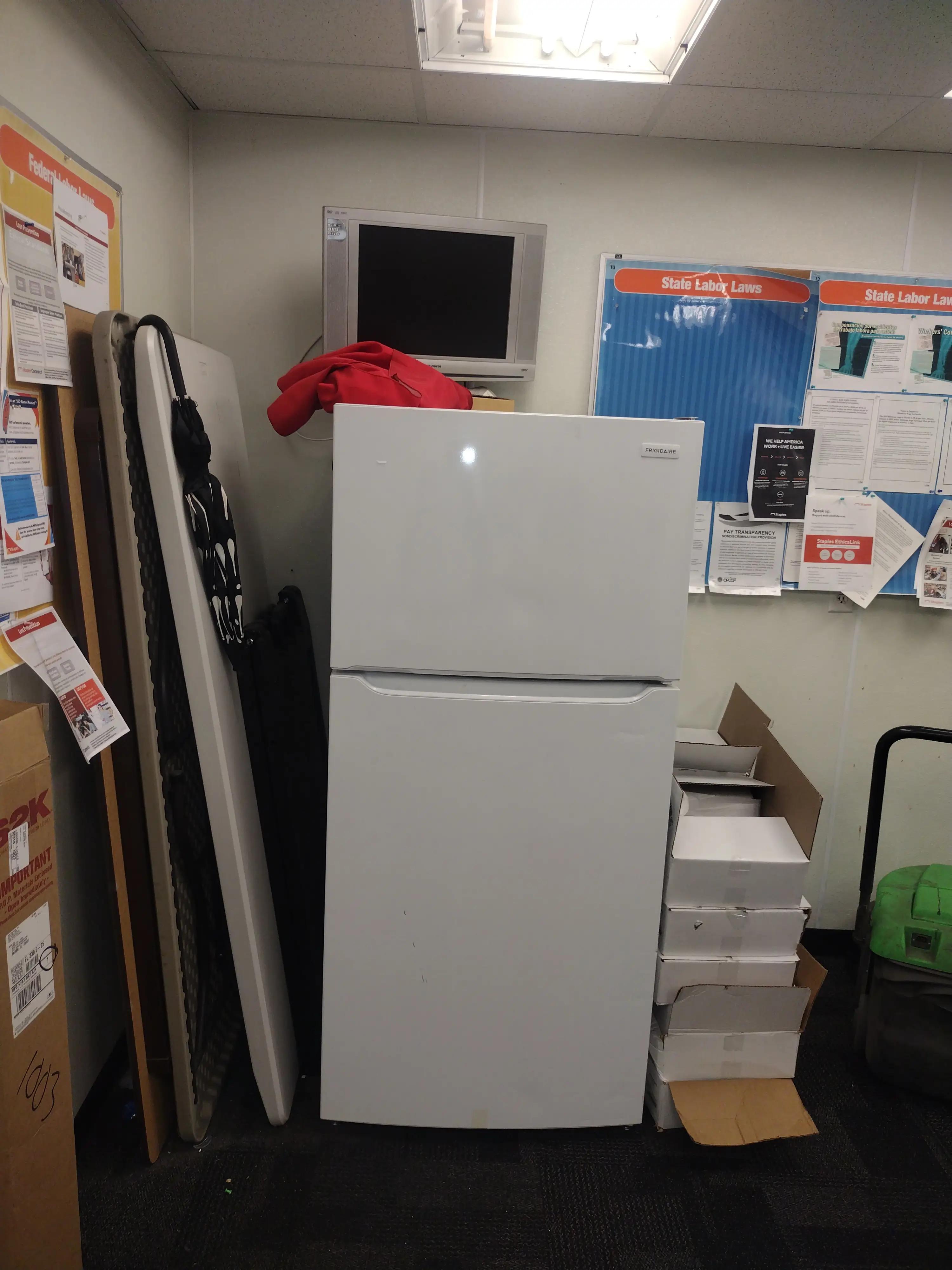 A workplace corner featuring a white refrigerator, a stack of boxes with a red jacket on top, a small TV, and various labor law posters on the walls.