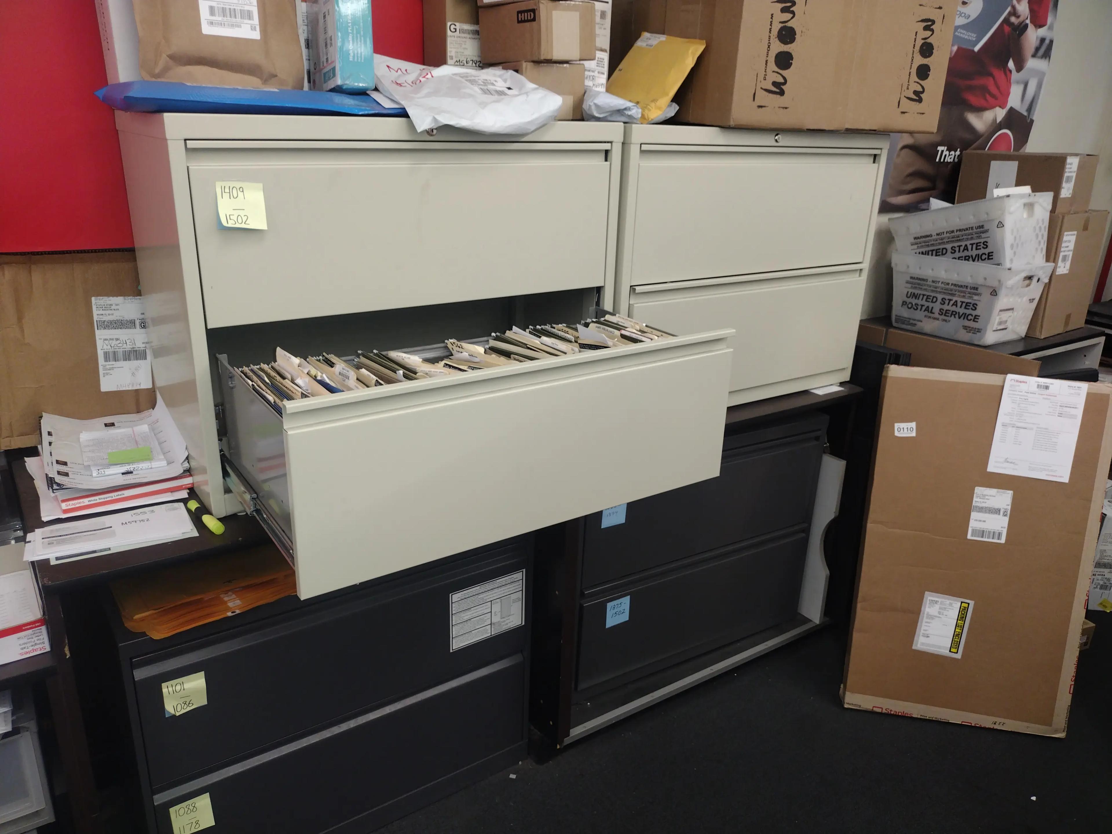 Office storage area with open filing cabinet drawers, various boxes, and postal service bins. Labels and documents are visible on the cabinets and boxes.