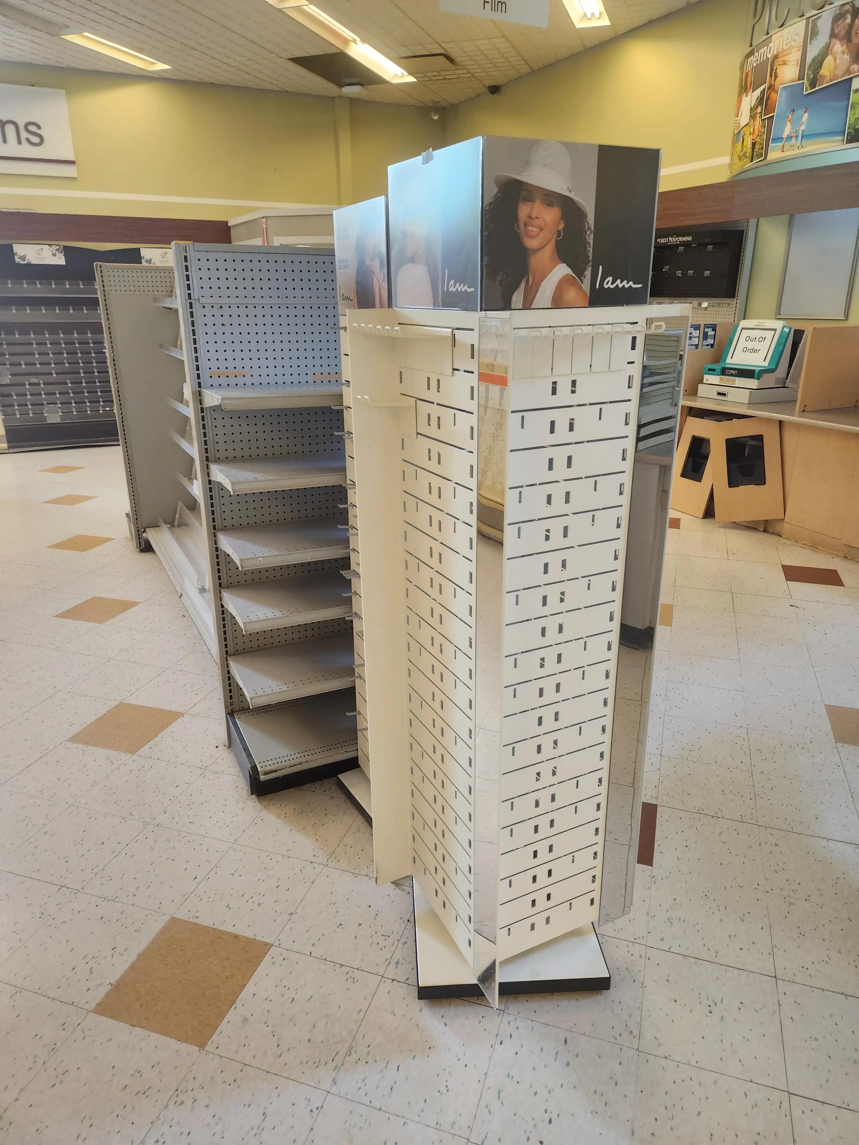 Empty retail display shelves and a rotating stand with a woman's image in a store setting. Signs for "Film" and "memories" are visible. The floor has a checkered pattern, and a sign reads "Out Of Order."