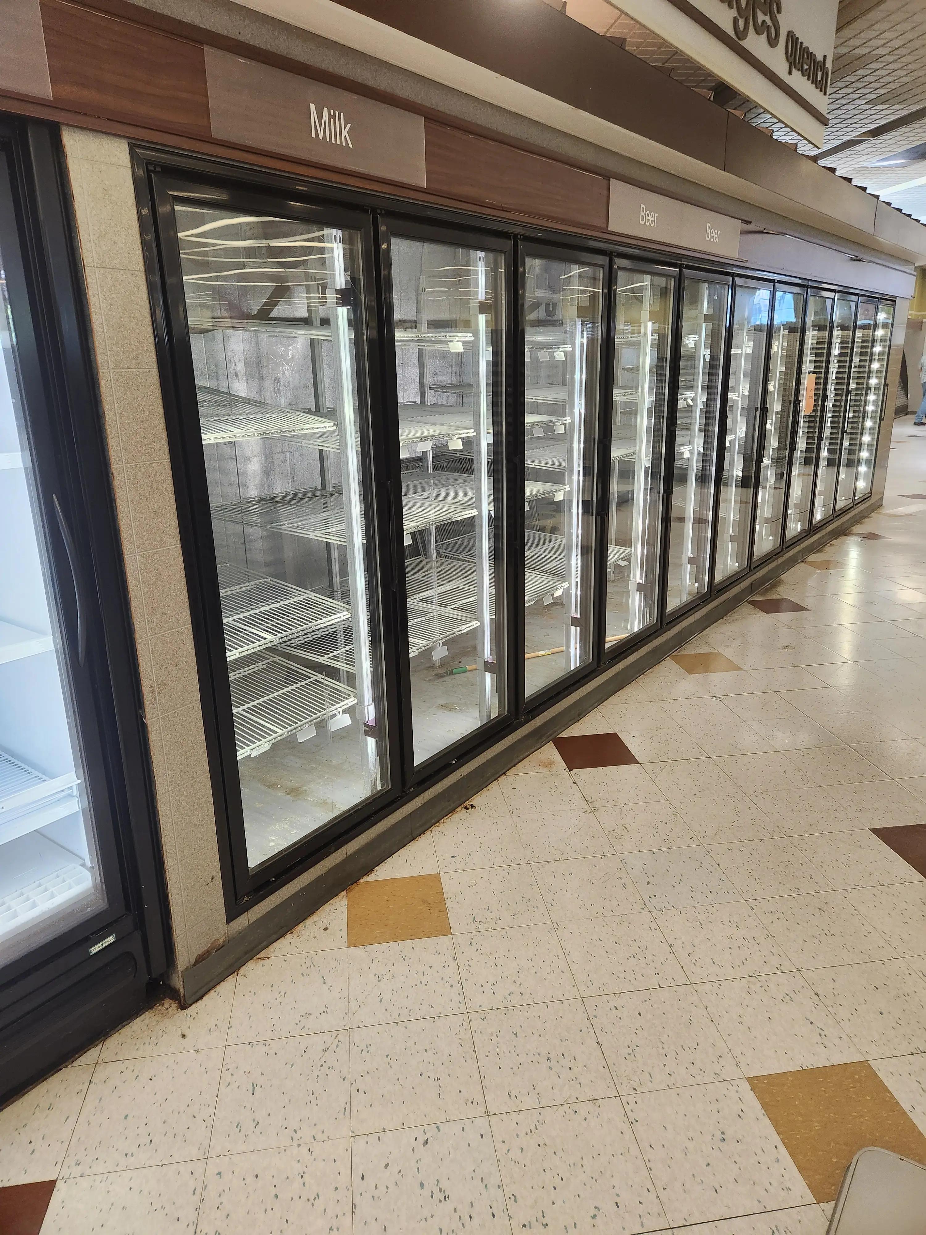 A row of empty refrigerated grocery store shelves labeled for milk and beer.