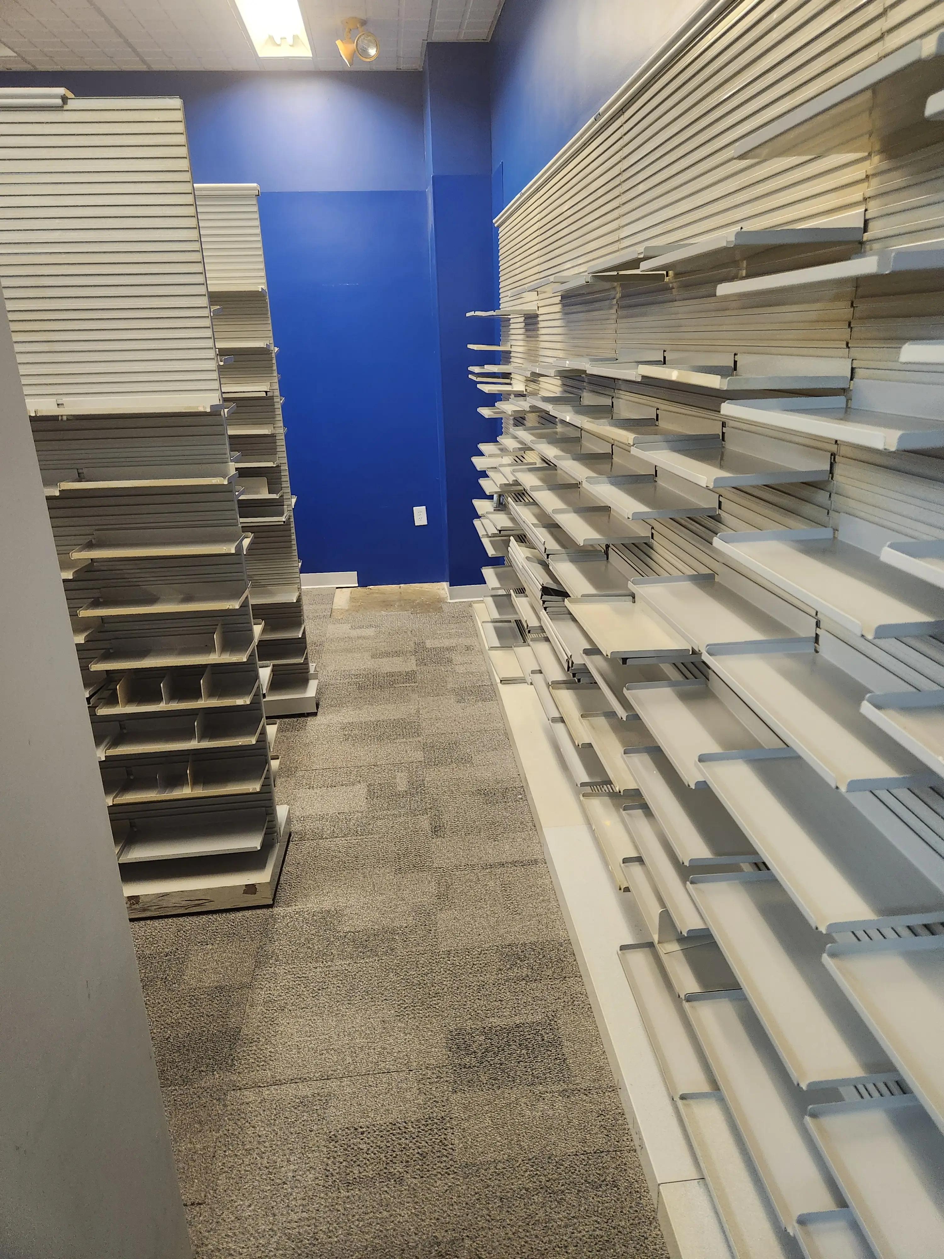 A room with blue walls, empty white retail shelving units on both sides, and a carpeted floor.