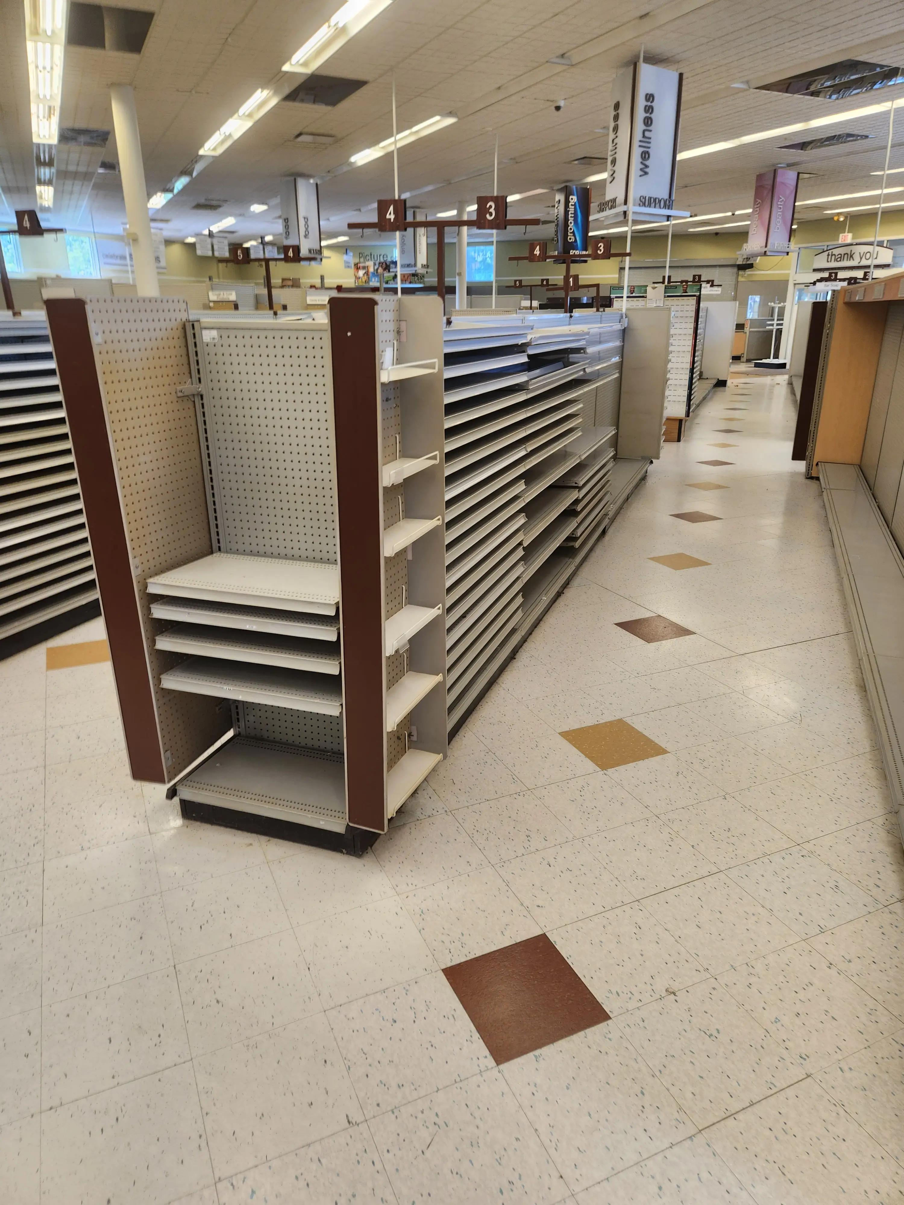 Empty store aisles with shelving units and overhead signs labeled "wellness," "food," "grooming," and "beauty." The floor features a pattern of white tiles with occasional brown and yellow tiles.