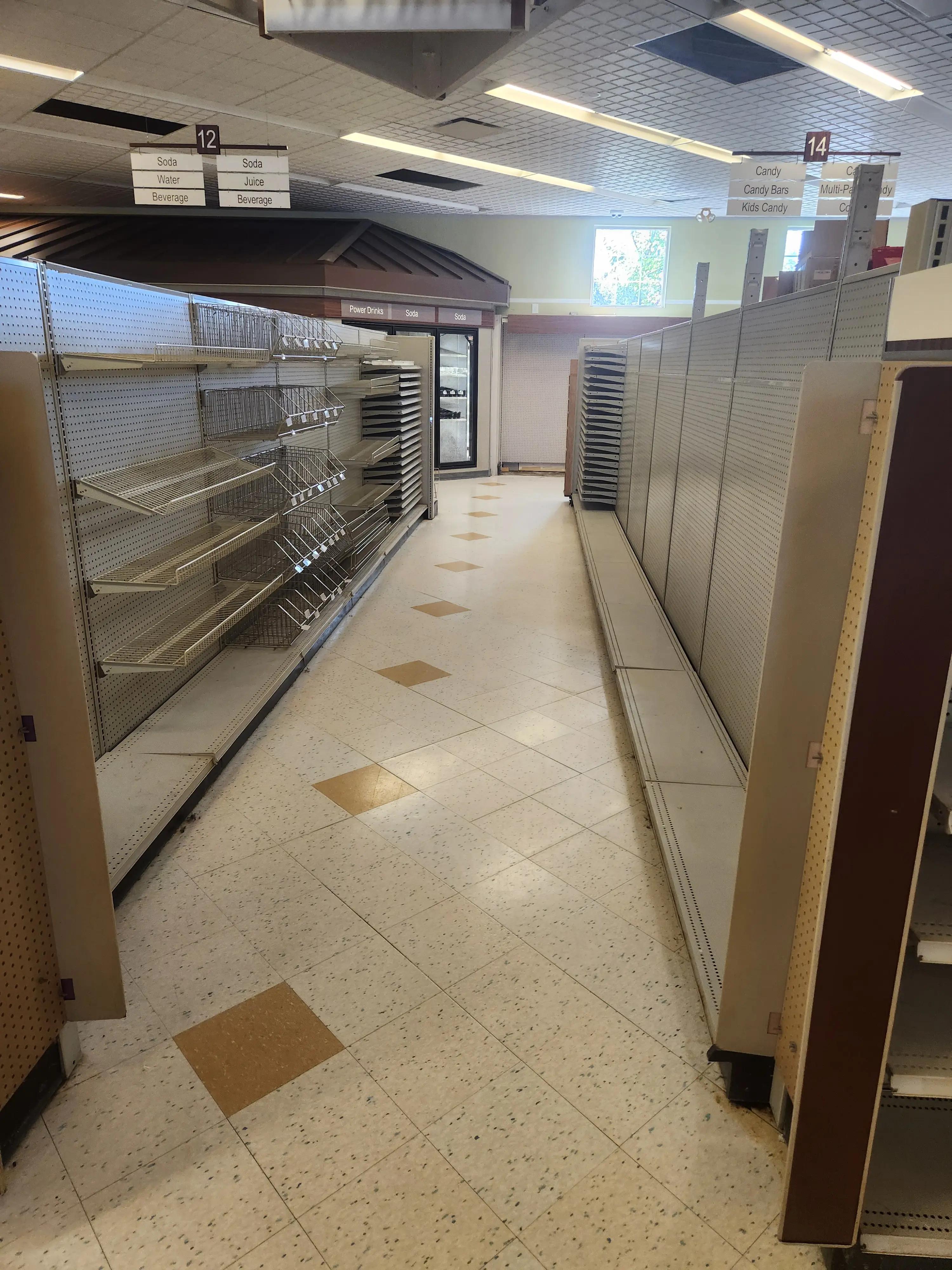 Empty grocery store aisles with metal shelves and overhead signs for soda, water, beverages, candy, and kids candy. The floor is tiled, and a window is visible in the background.