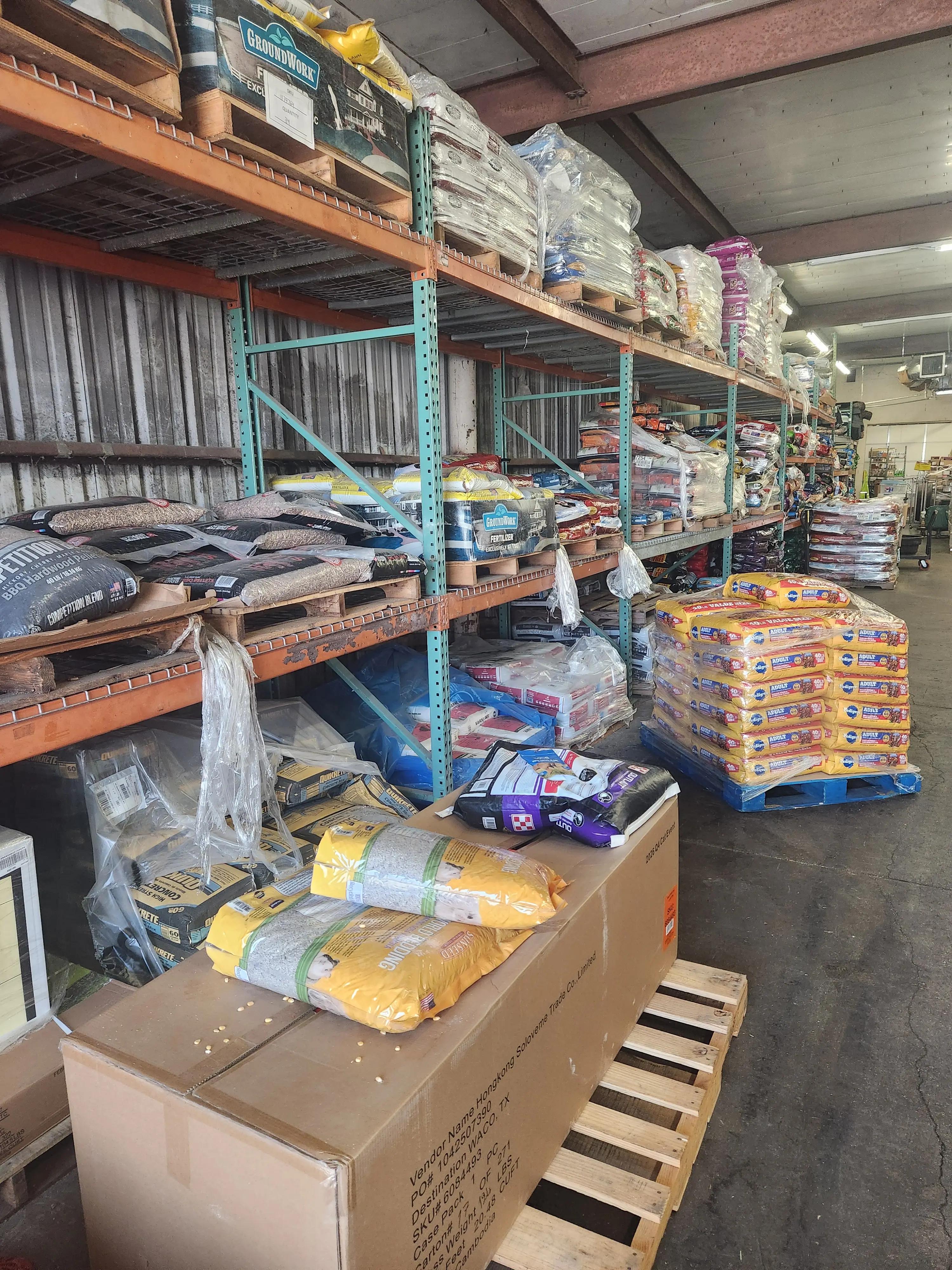 Warehouse shelves filled with bags of lawn care products, fertilizers, and BBQ hardwood pellets from brands like Groundwork and Quikrete. The industrial storage area features metal shelving and stacked pallets.