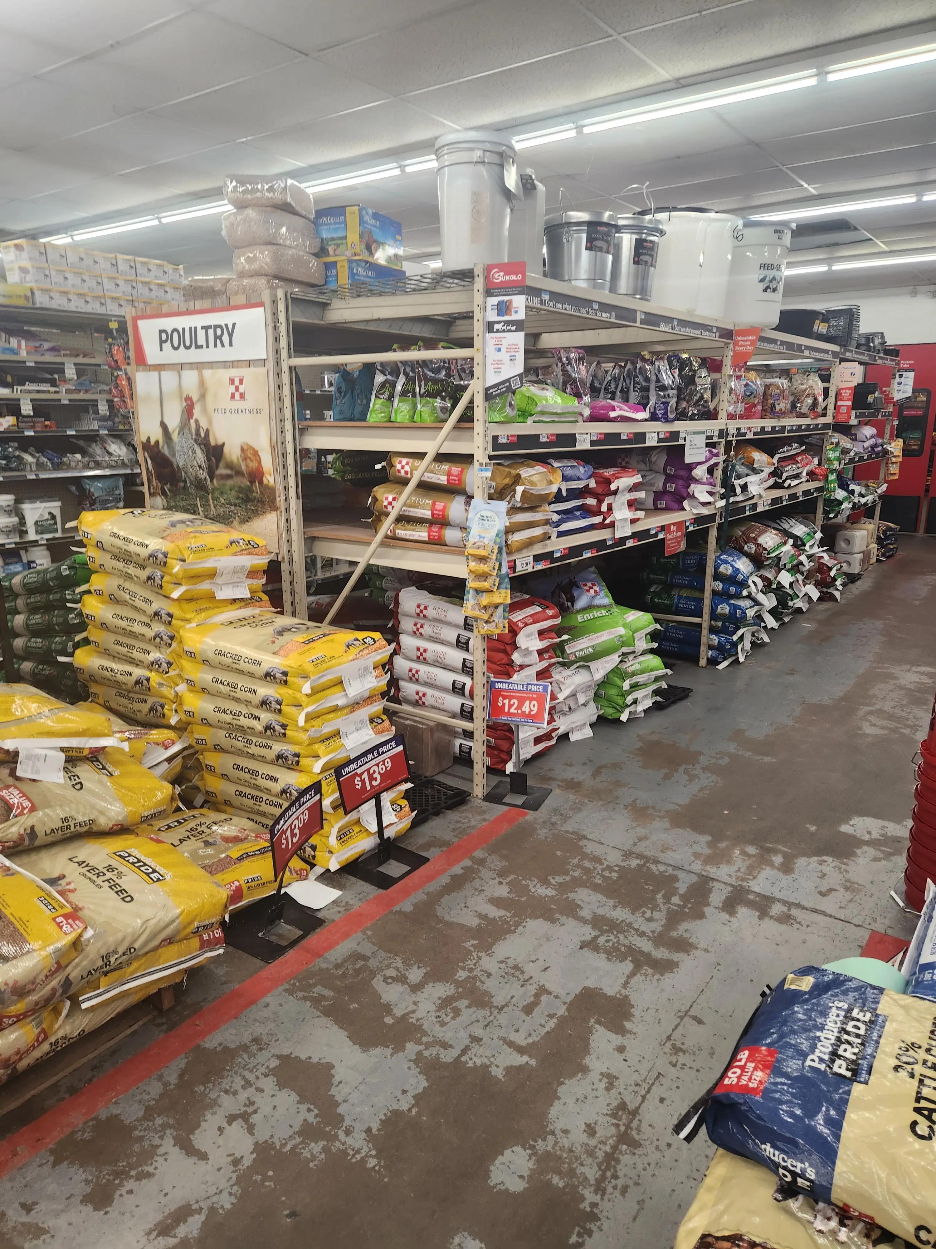 Store aisle with shelves stocked with various animal feed bags, including poultry and cracked corn. Signs display prices like $13.99 and $12.49. Buckets and containers are on the top shelf.