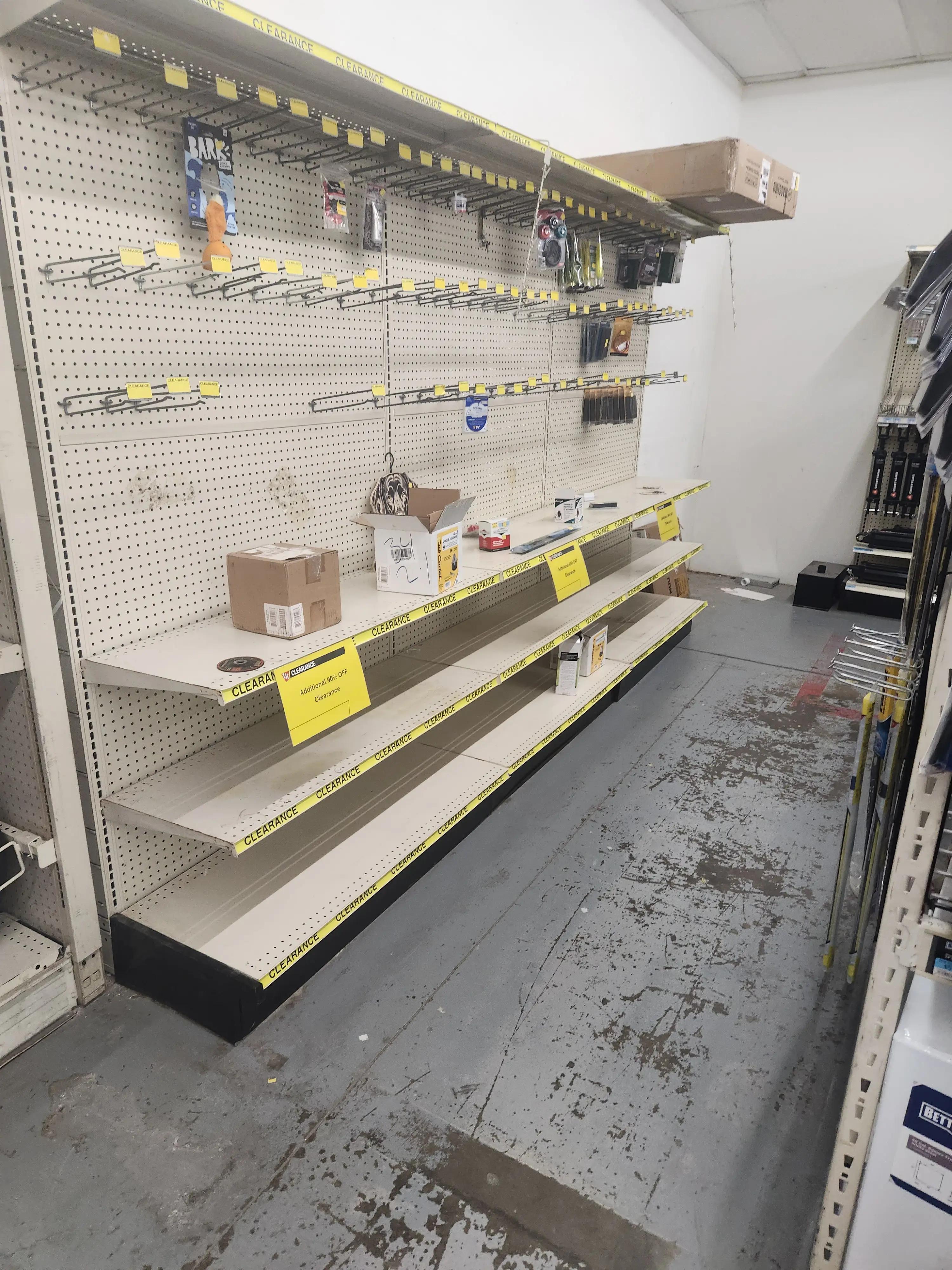 Alt-text: Image of mostly empty store shelves with yellow clearance signs and tape, indicating a significant sale. A few remaining items, including boxes and small products, are visible. The floor shows signs of wear.