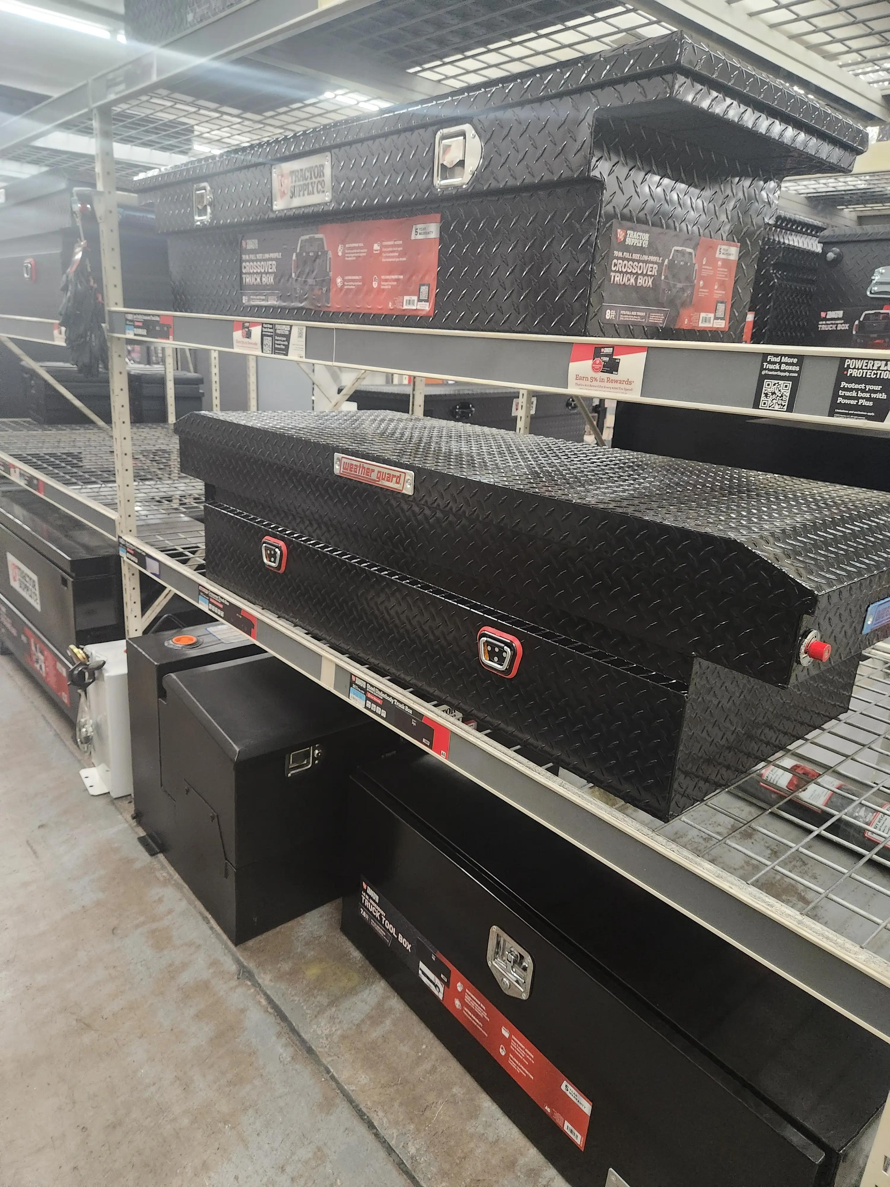 Shelves displaying various black truck tool boxes at a store, including crossover and underbody models, with branding from Tractor Supply Co and Weather Guard.