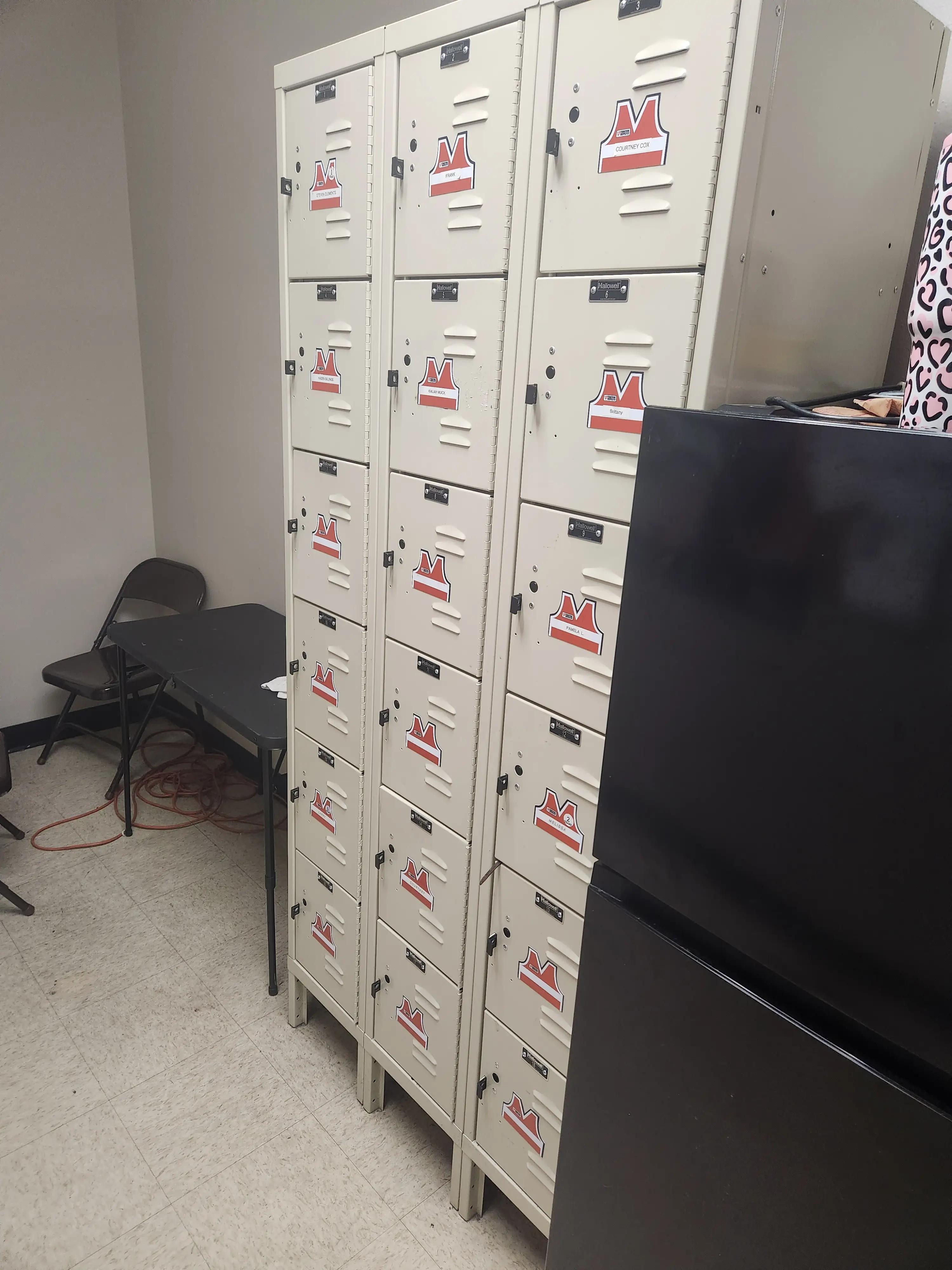 A row of beige lockers with red and white Mid America logos, each labeled with different names, next to a black refrigerator and a folding table with chairs in a room.