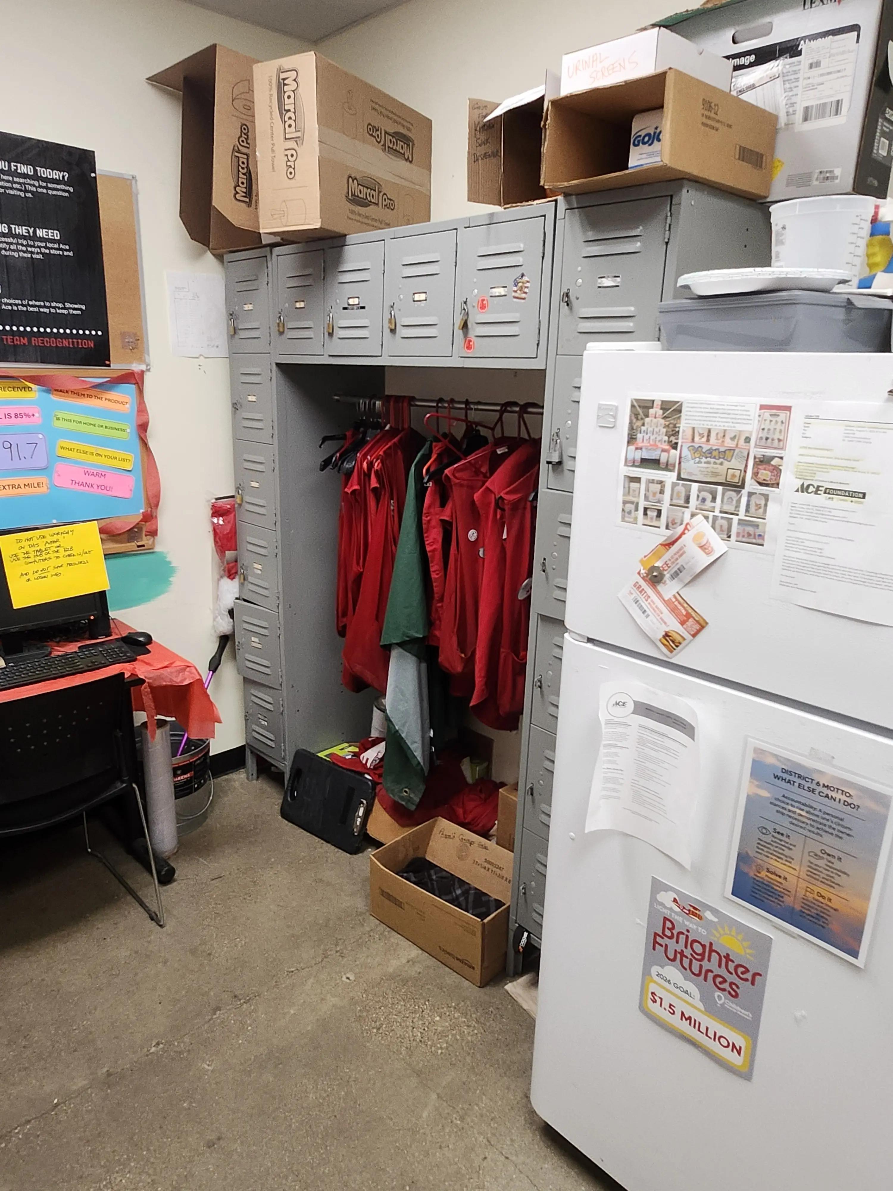 Storage room with lockers, red jackets hanging, boxes labeled "Marcal Pro" on top, a refrigerator with papers and a "Brighter Futures" poster, and a desk with a computer on the left.