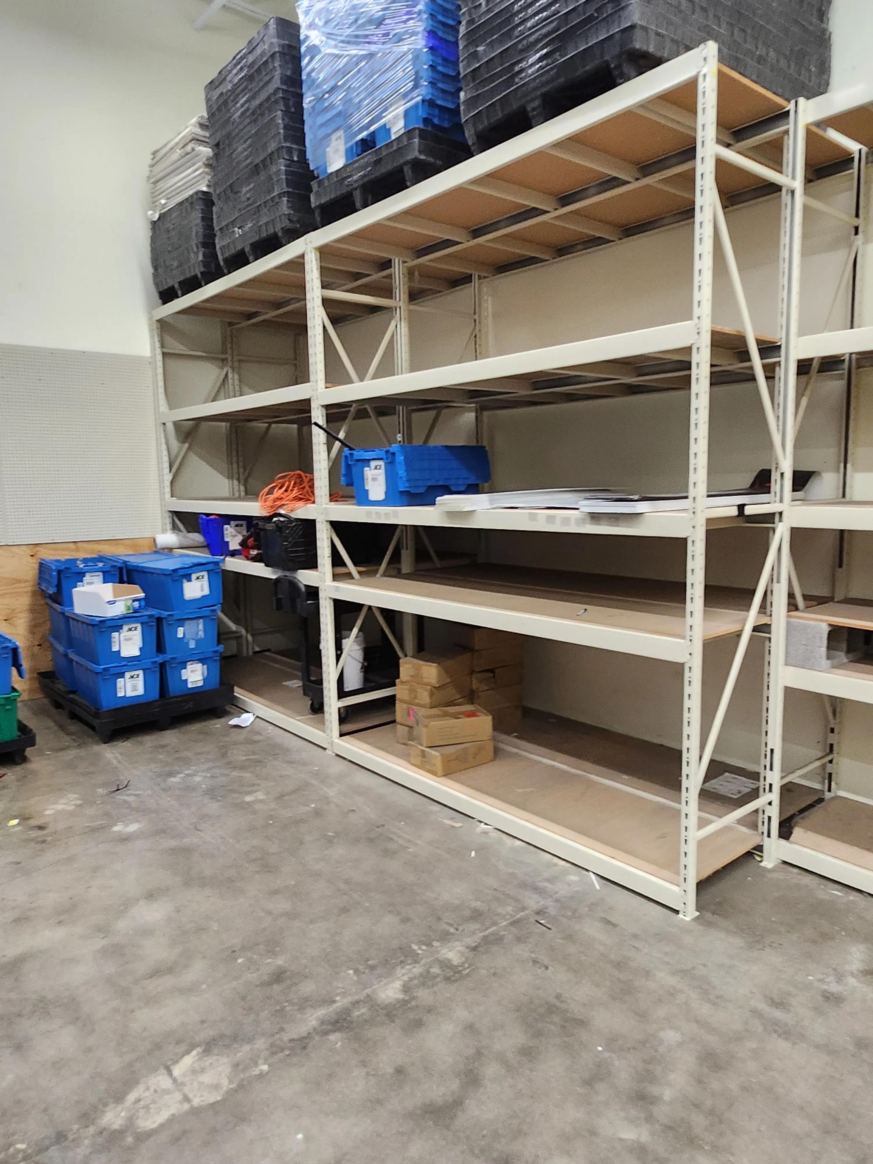 Industrial storage shelves filled with blue plastic bins, cardboard boxes, and stacked pallets in a warehouse environment.