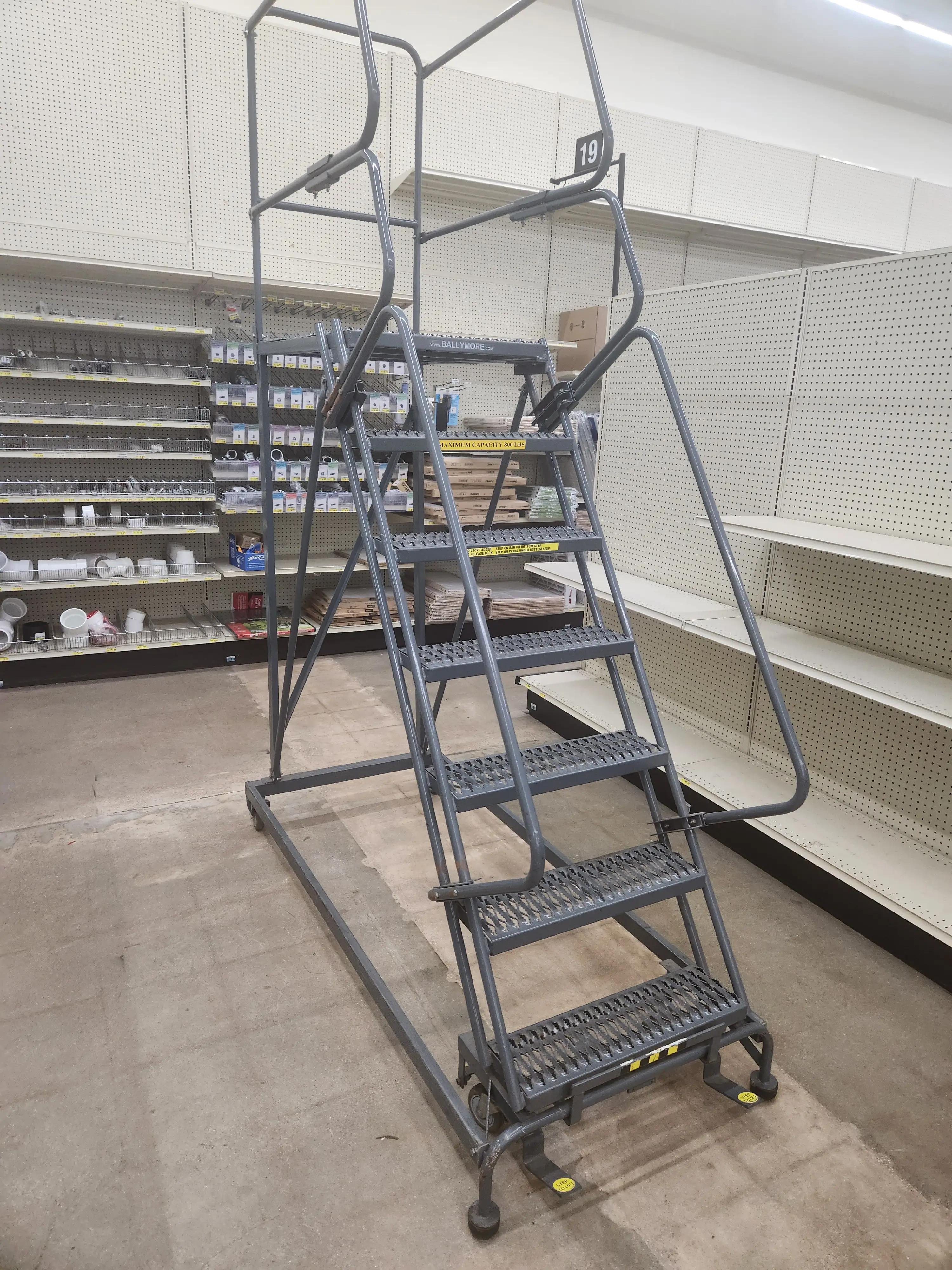 A gray rolling ladder with handrails and steps is positioned in a store aisle, surrounded by shelves with various items. The ladder has a maximum capacity of 800 lbs and features a locking mechanism.
