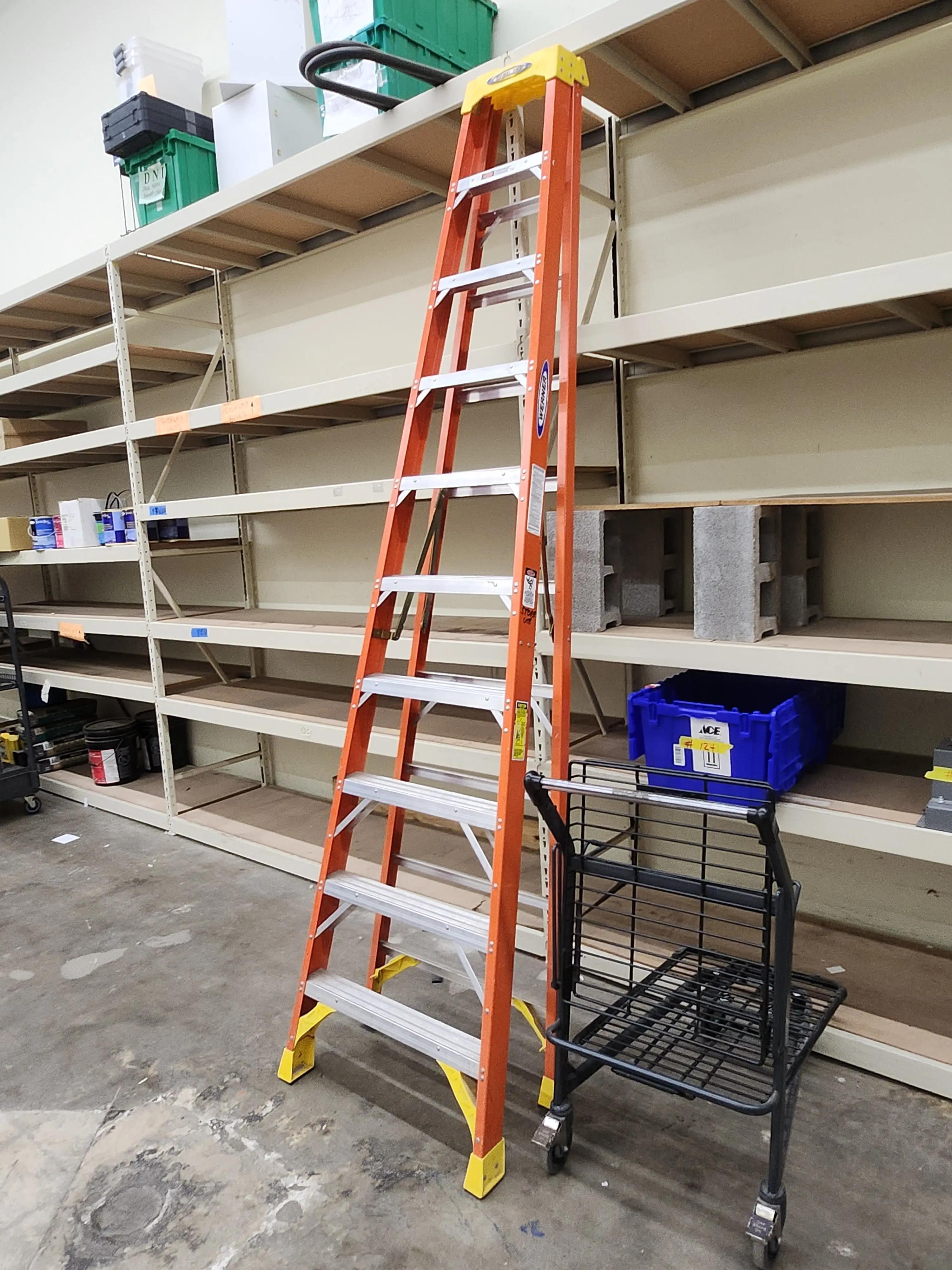 Orange ladder leaning against empty metal shelving in a storage area, with a cart and a blue storage bin nearby.